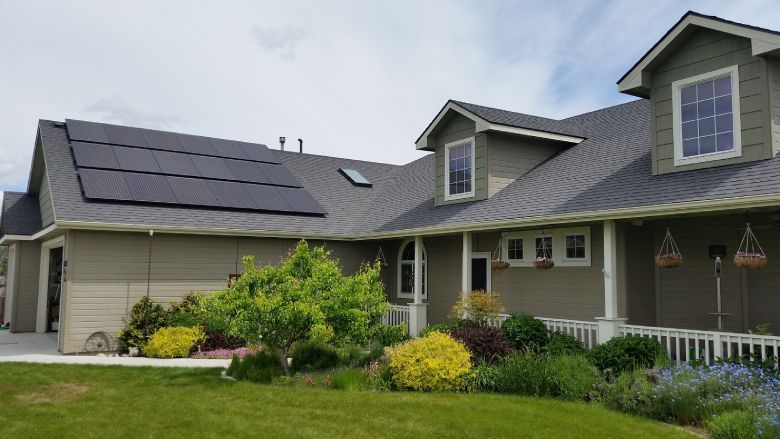 Solar panels on a green house roof with a well-manicured lawn and flower garden. Blue sky.