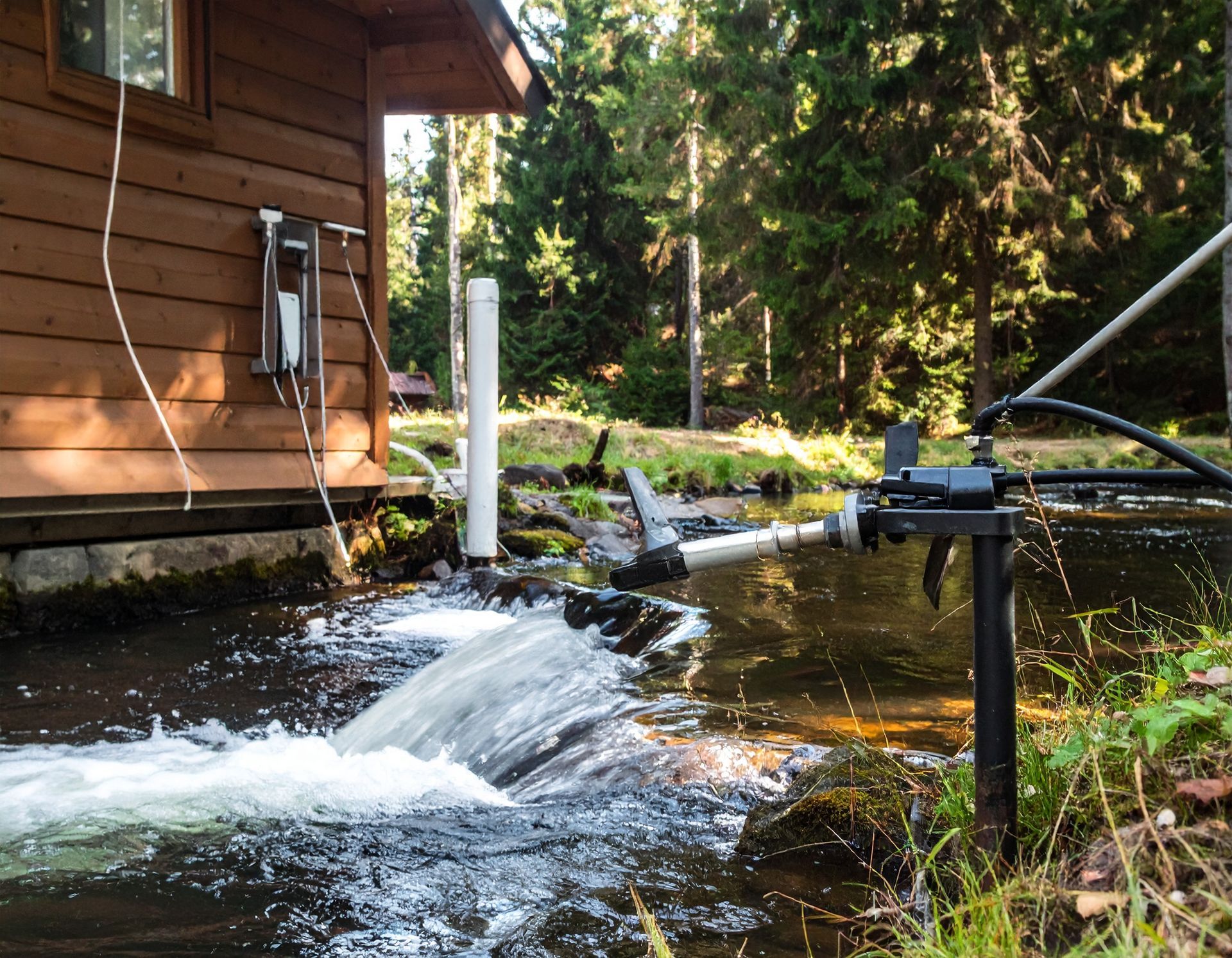 Small scale micro-hydro turbine system generating electricity from a stream next to a wooden cabin in a forest.