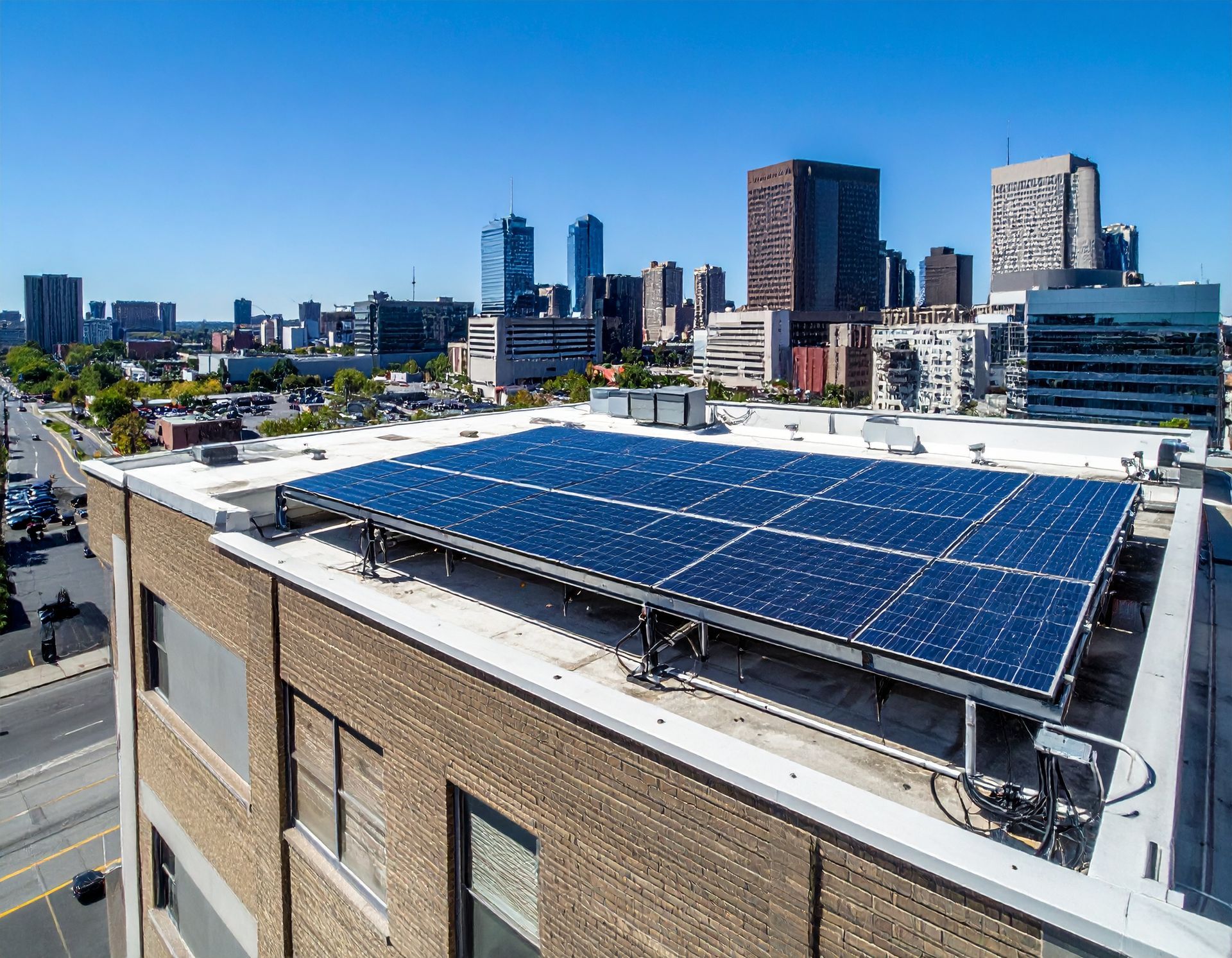 A solar panel array sits on the flat roof of a brick building overlooking an urban city skyline on a clear, sunny day.