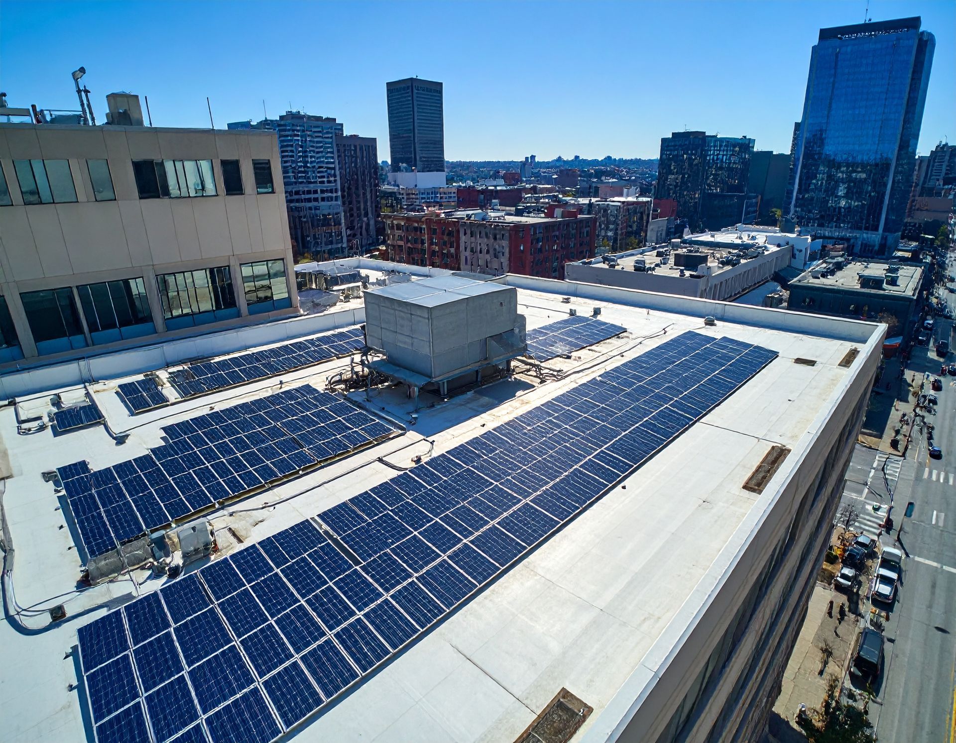 Aerial view of a city rooftop covered in solar panels with tall buildings in the background under a bright blue sky.