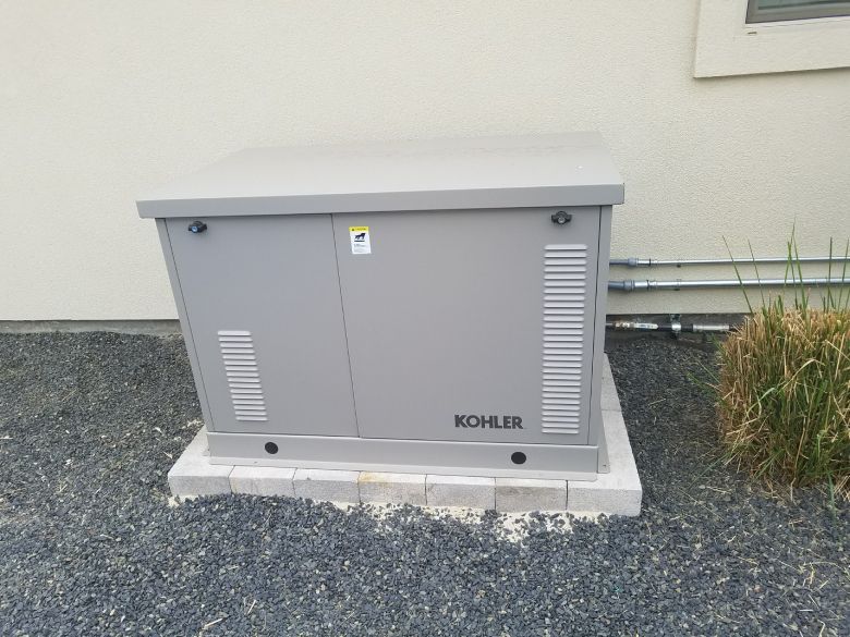 Gray Kohler home generator on a concrete base, next to a beige building with gravel ground.