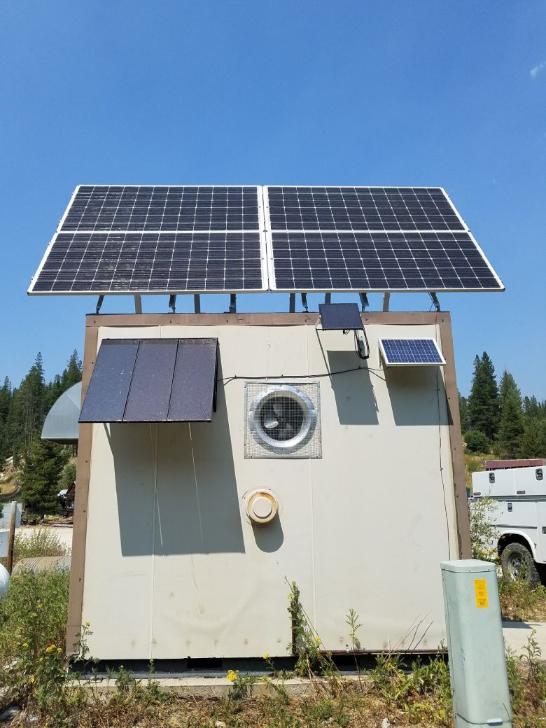 Solar panels mounted on a small building with an open vent and two additional small panels, set against a blue sky.