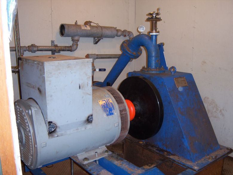 Blue and gray generator equipment in a utility room; pipes and a gauge are attached.