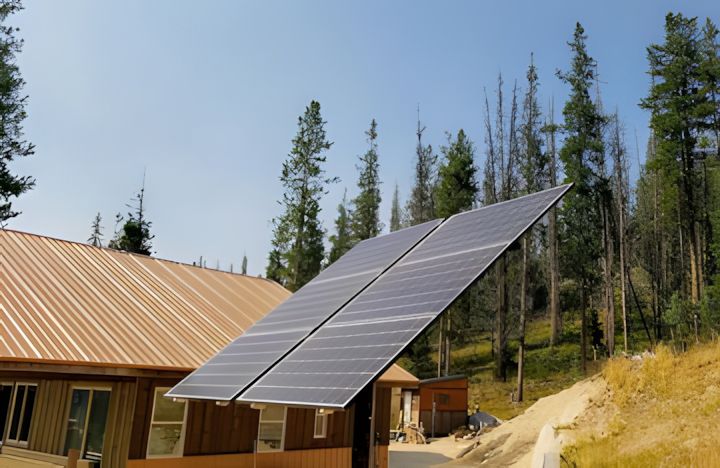 Solar panels angled toward the sky, mounted near a wood-sided building, with trees in the background.