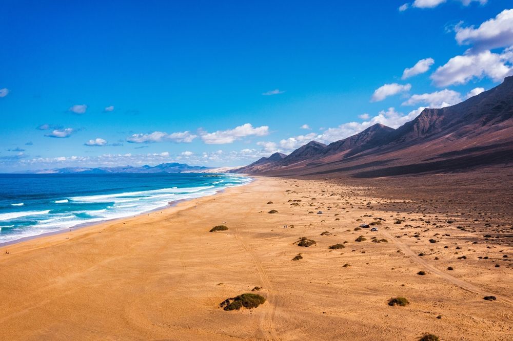 Sandy beach along blue ocean with mountains and bright blue sky.