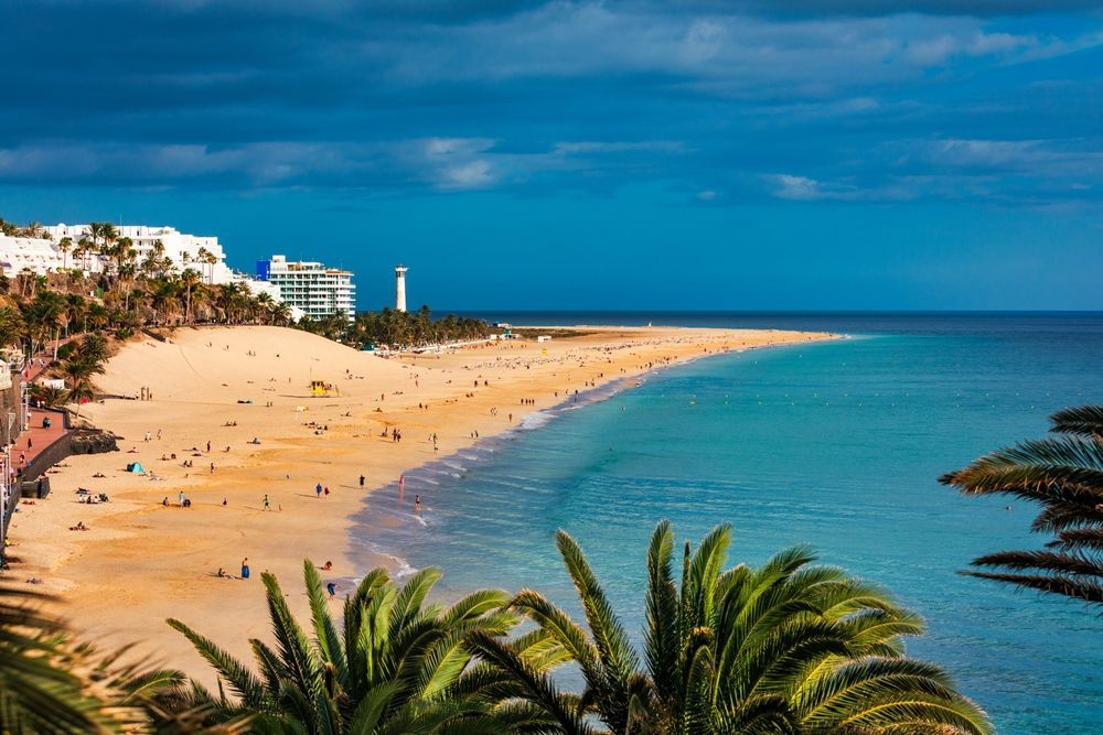 Sandy beach with turquoise water, palm trees, and buildings under a bright blue sky.