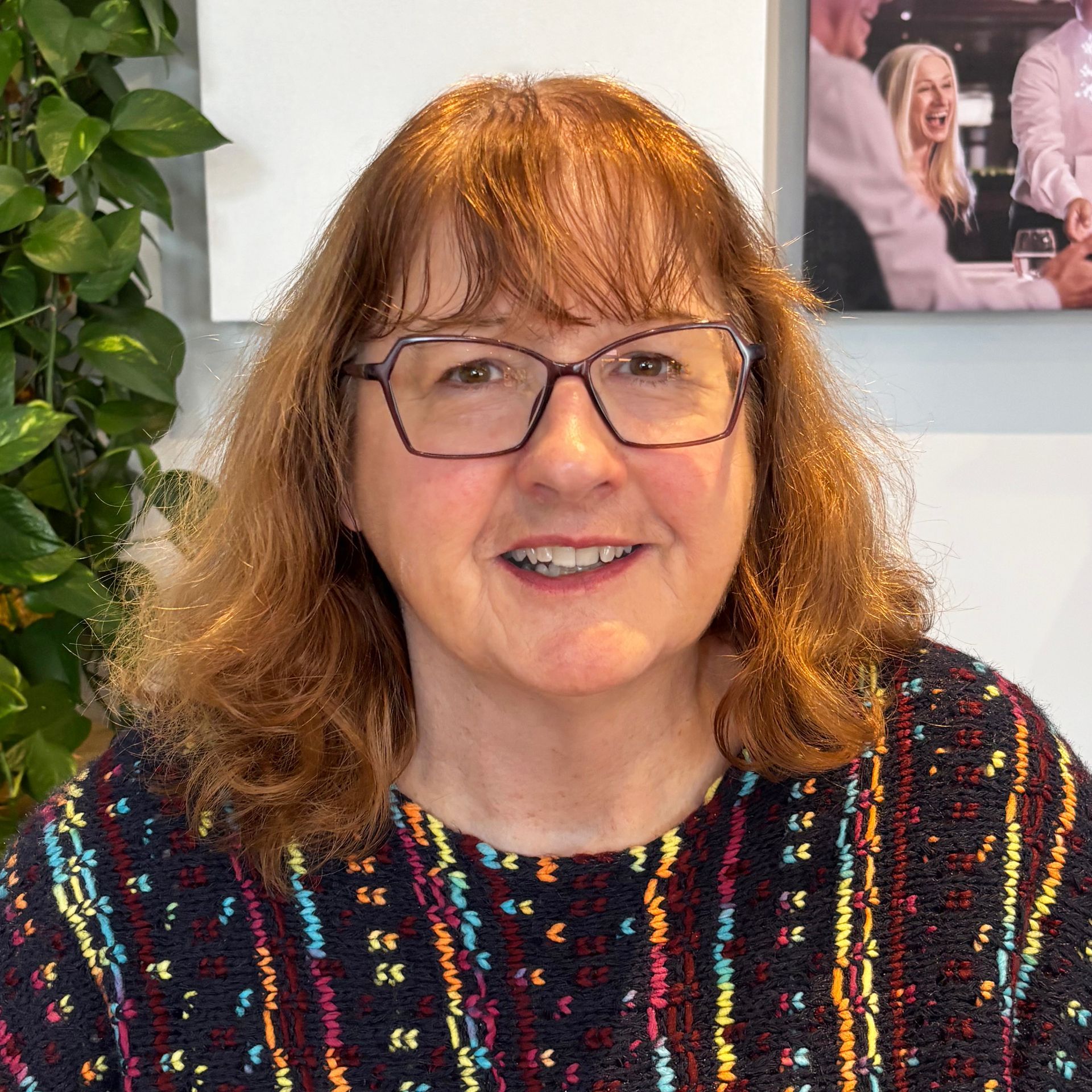 Woman with glasses and auburn hair smiles, wearing a patterned top, indoor setting with a photo and plants.