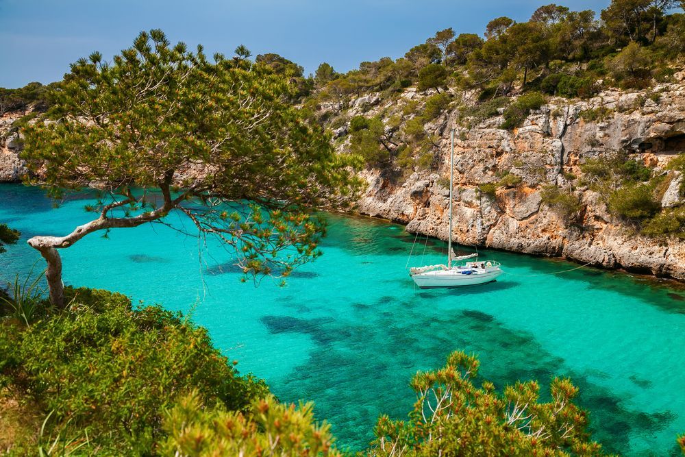 Bright turquoise bay with white sailboat, rocky cliffs, and green trees under a blue sky.