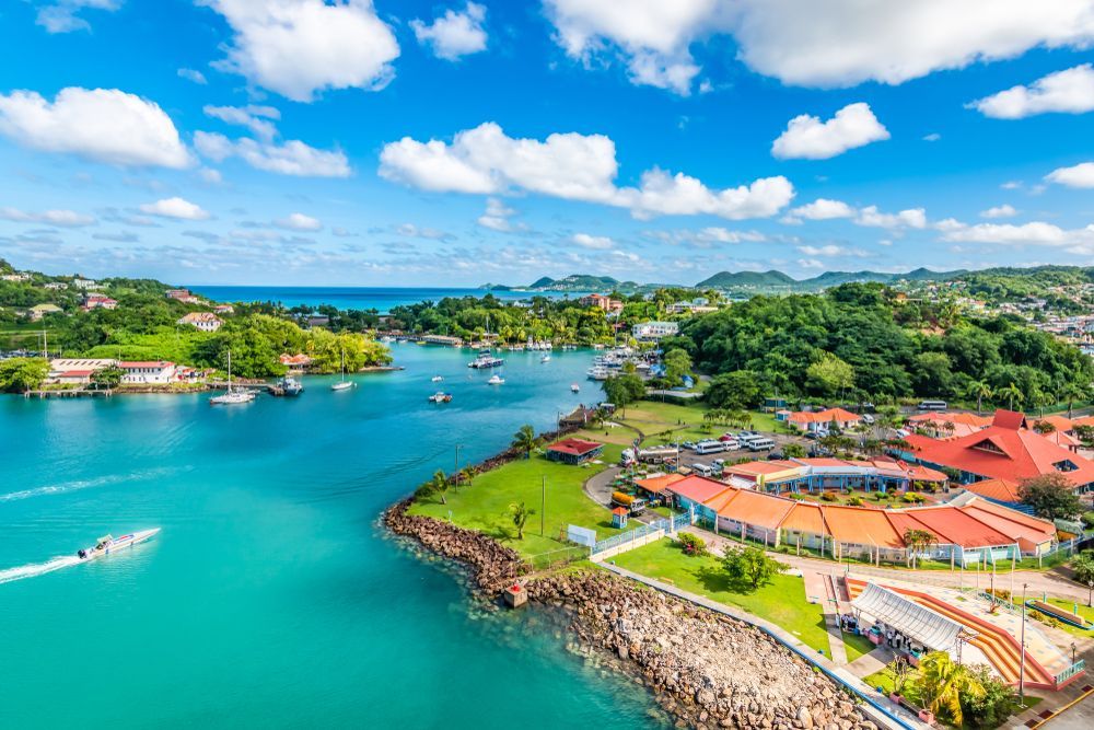 Turquoise bay with boats, surrounded by green hills, red-roofed buildings, and a partly cloudy blue sky.