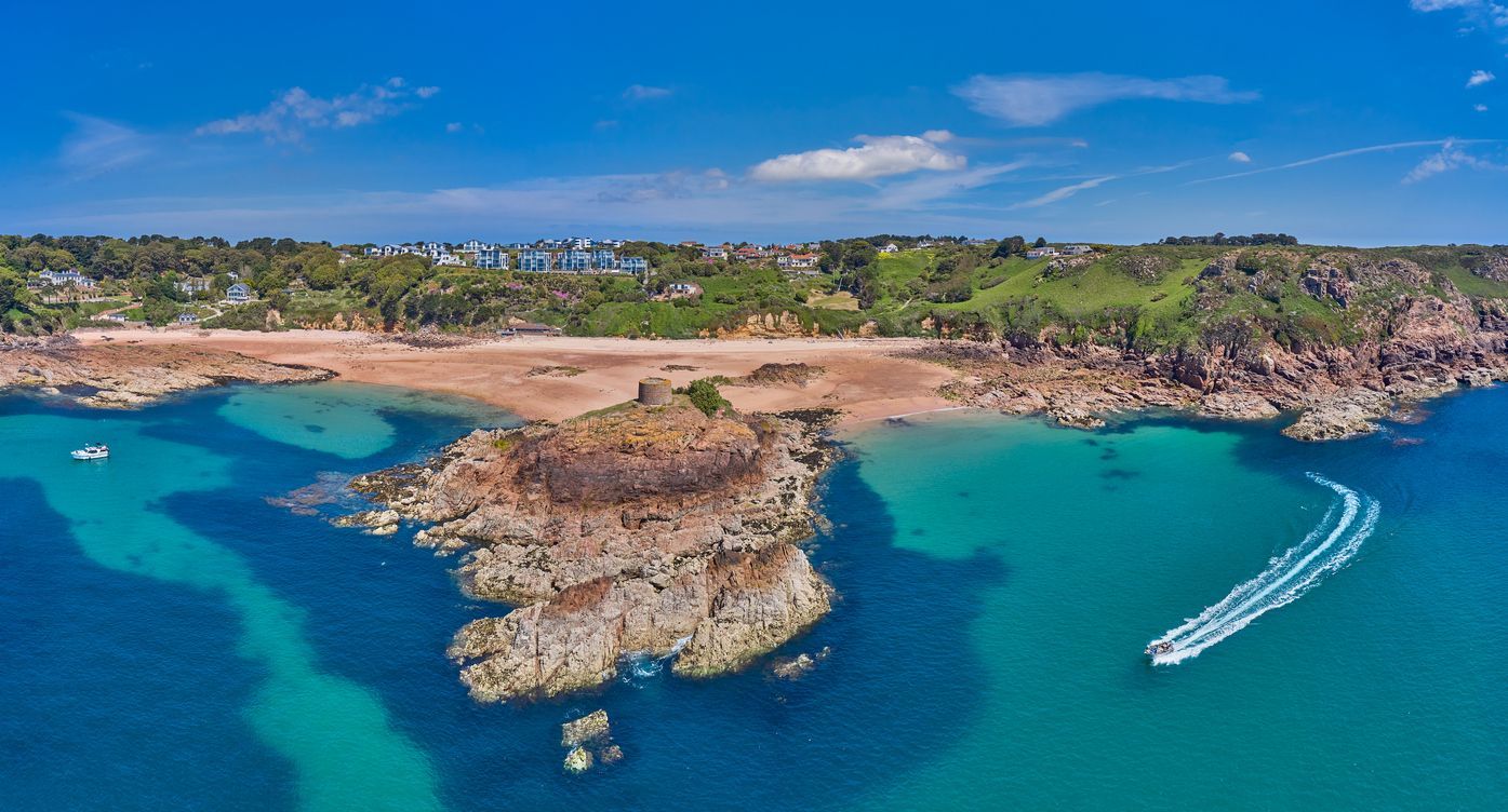Aerial view of a sandy beach, turquoise water, rocky cliffs, and a boat with white wake under a bright blue sky.