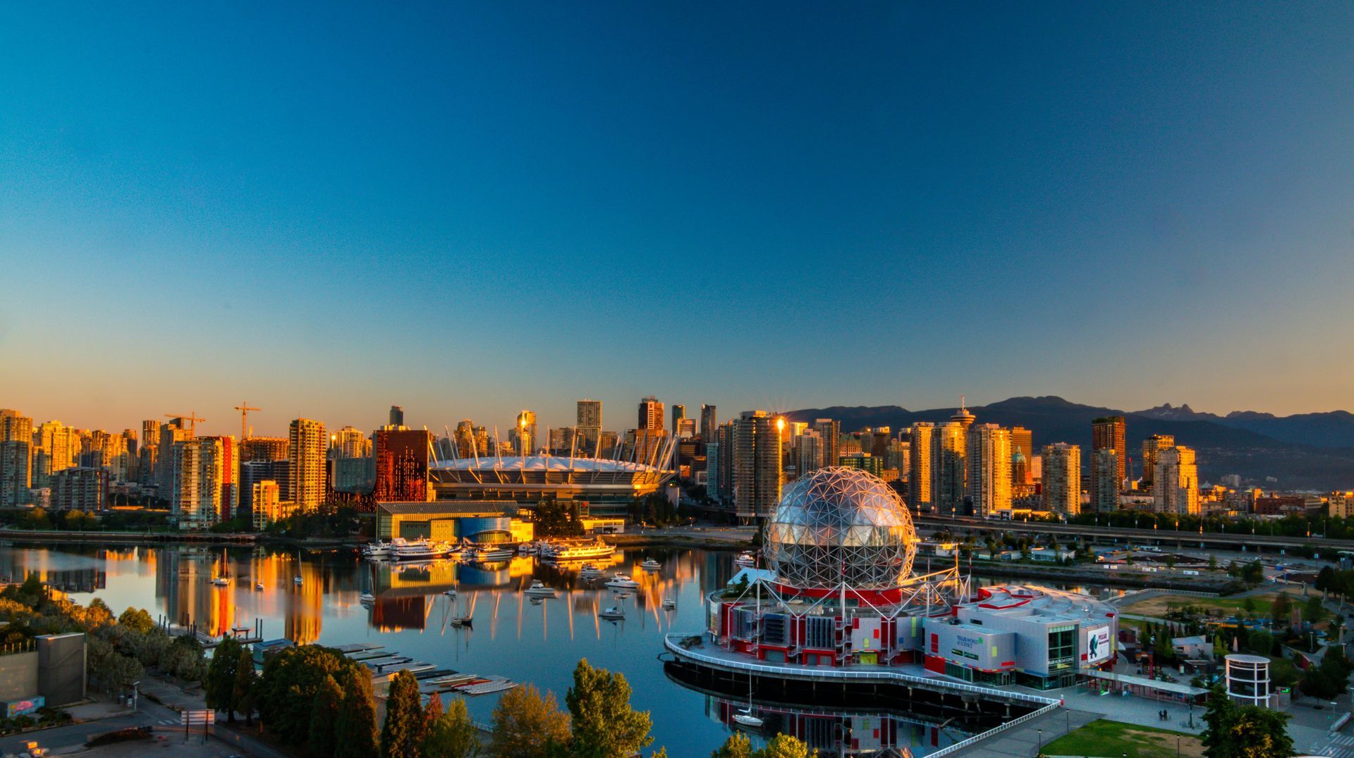 City skyline reflecting in water with a large spherical building. Golden hour lighting illuminates buildings and sky.