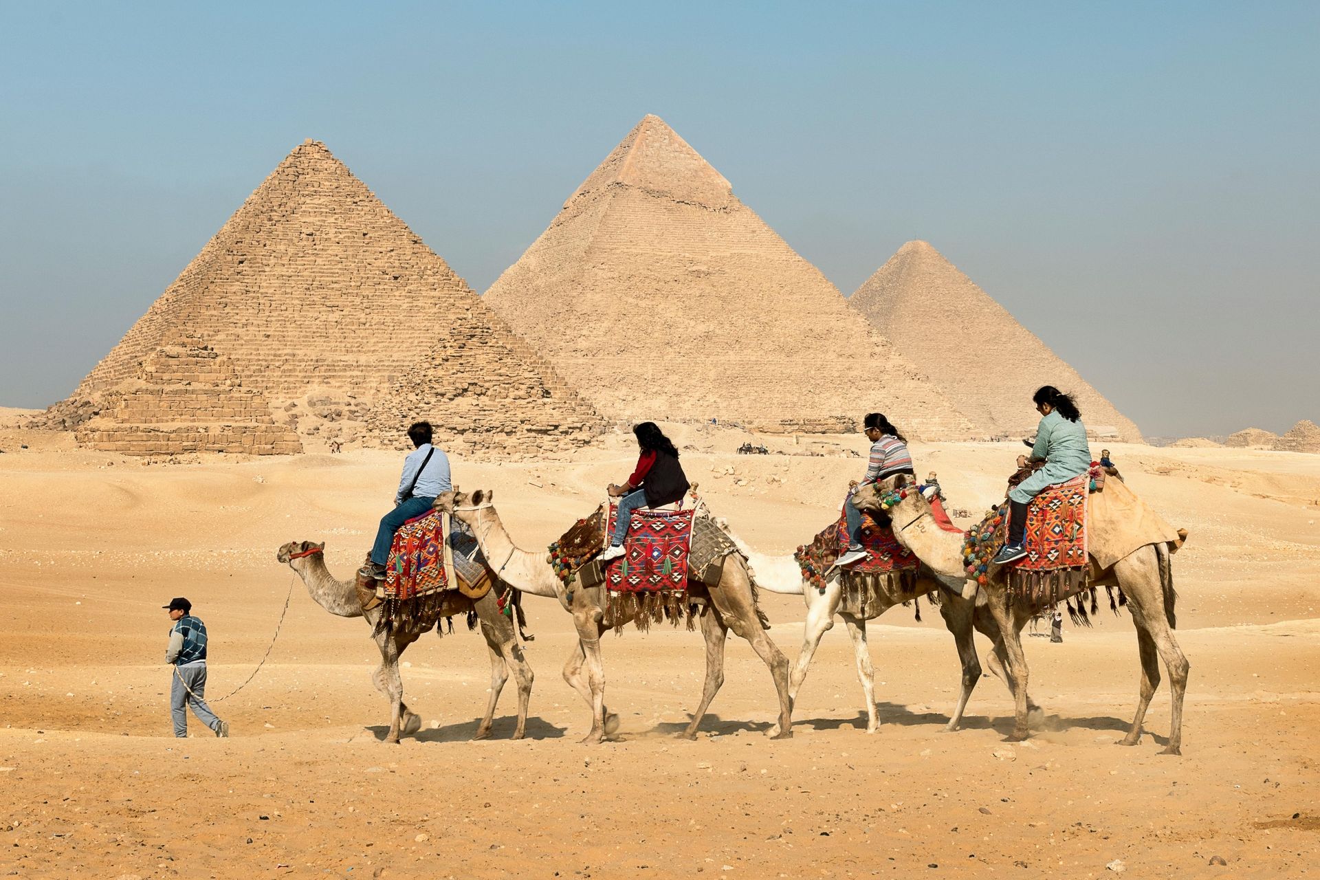 People riding camels in front of the pyramids in Egypt; sunny, desert landscape.