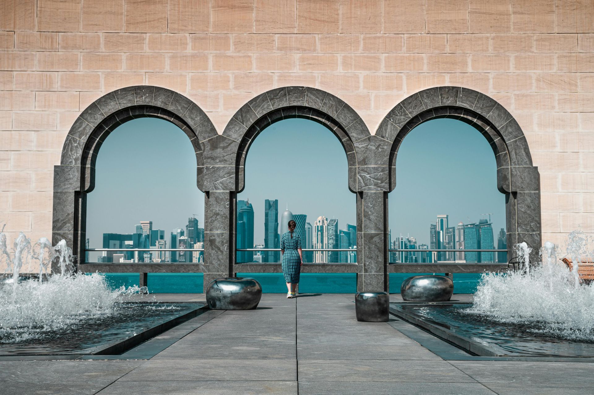 Person stands between arched windows, city skyline backdrop. Water fountains on either side, beige stone.