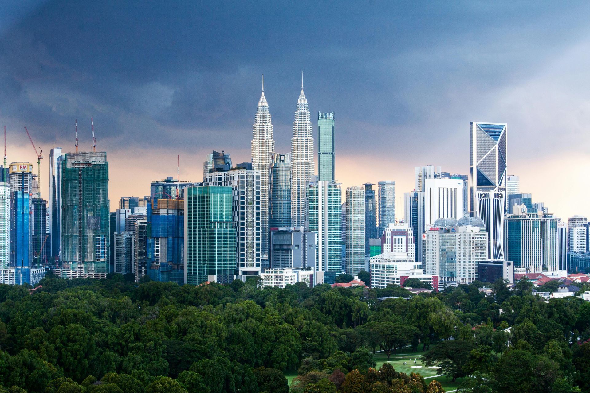 City skyline with Petronas Towers against a cloudy sky, green trees in the foreground.