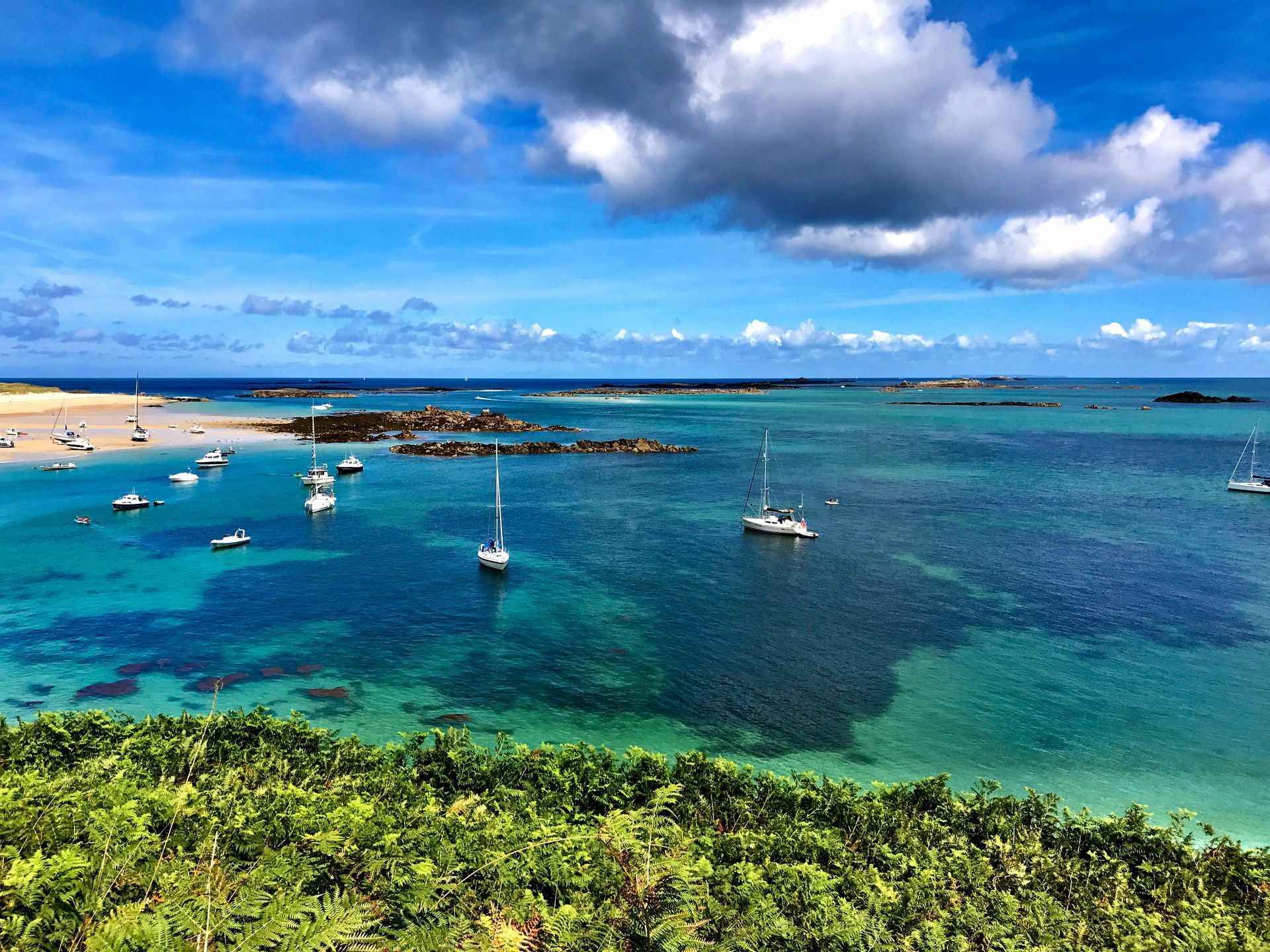 Bright blue ocean with white sailboats, rocky islands, and a green, sunny hillside.