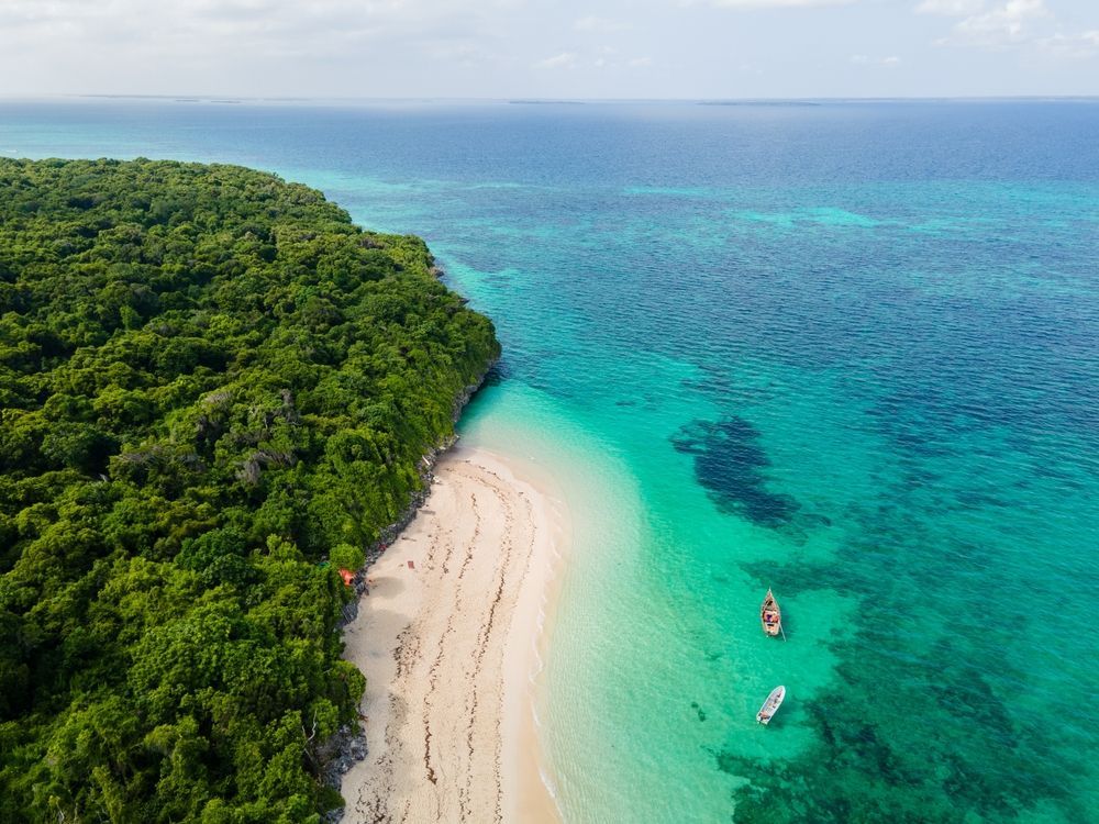 Aerial view of a white sand beach bordered by green forest and turquoise water; two small boats float offshore.
