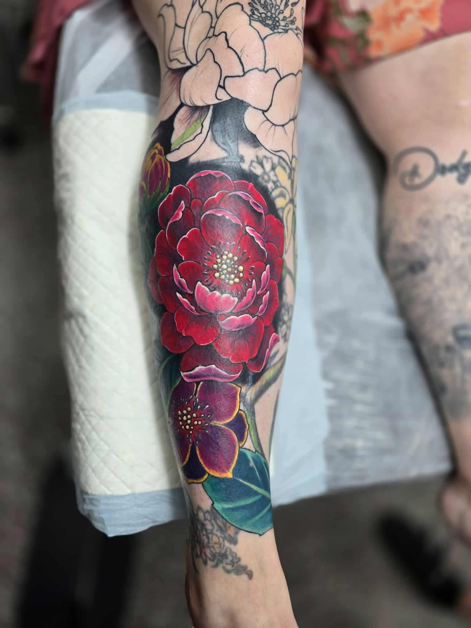 Colorful peony tattoo on a leg; red and purple blooms with green leaves; detailed, artistic style.