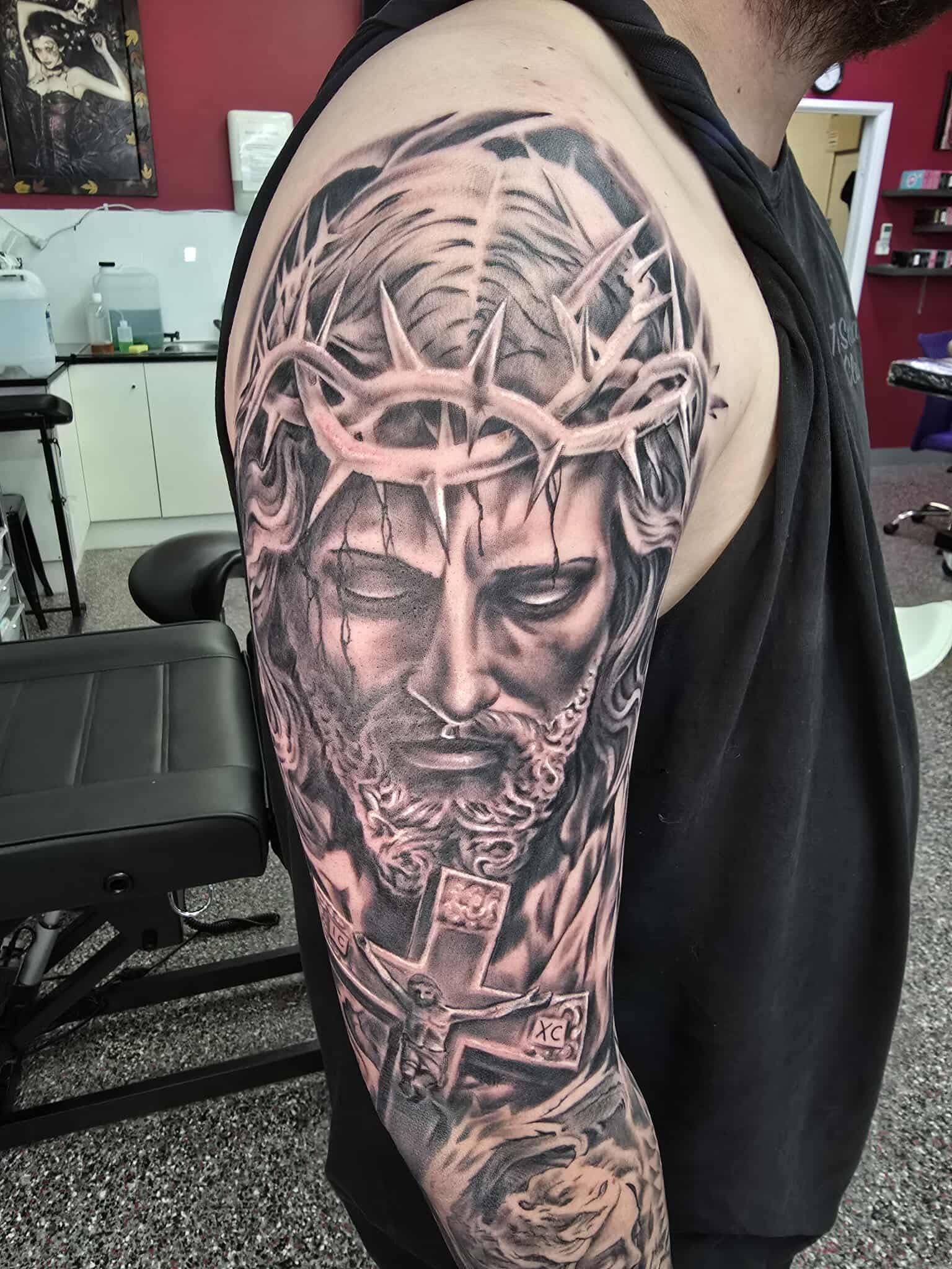 A man's arm tattoo of Jesus wearing a crown of thorns and a cross; black and gray ink.