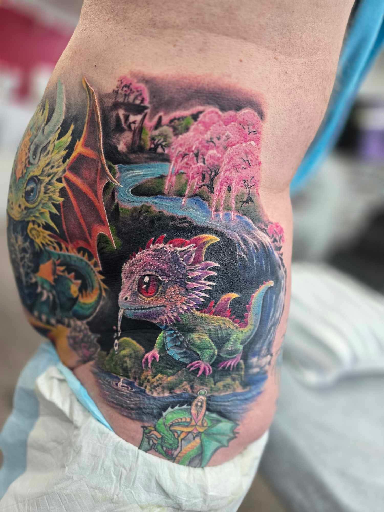 Dragon tattoo: baby dragon near a waterfall, and a larger dragon in a colorful landscape.