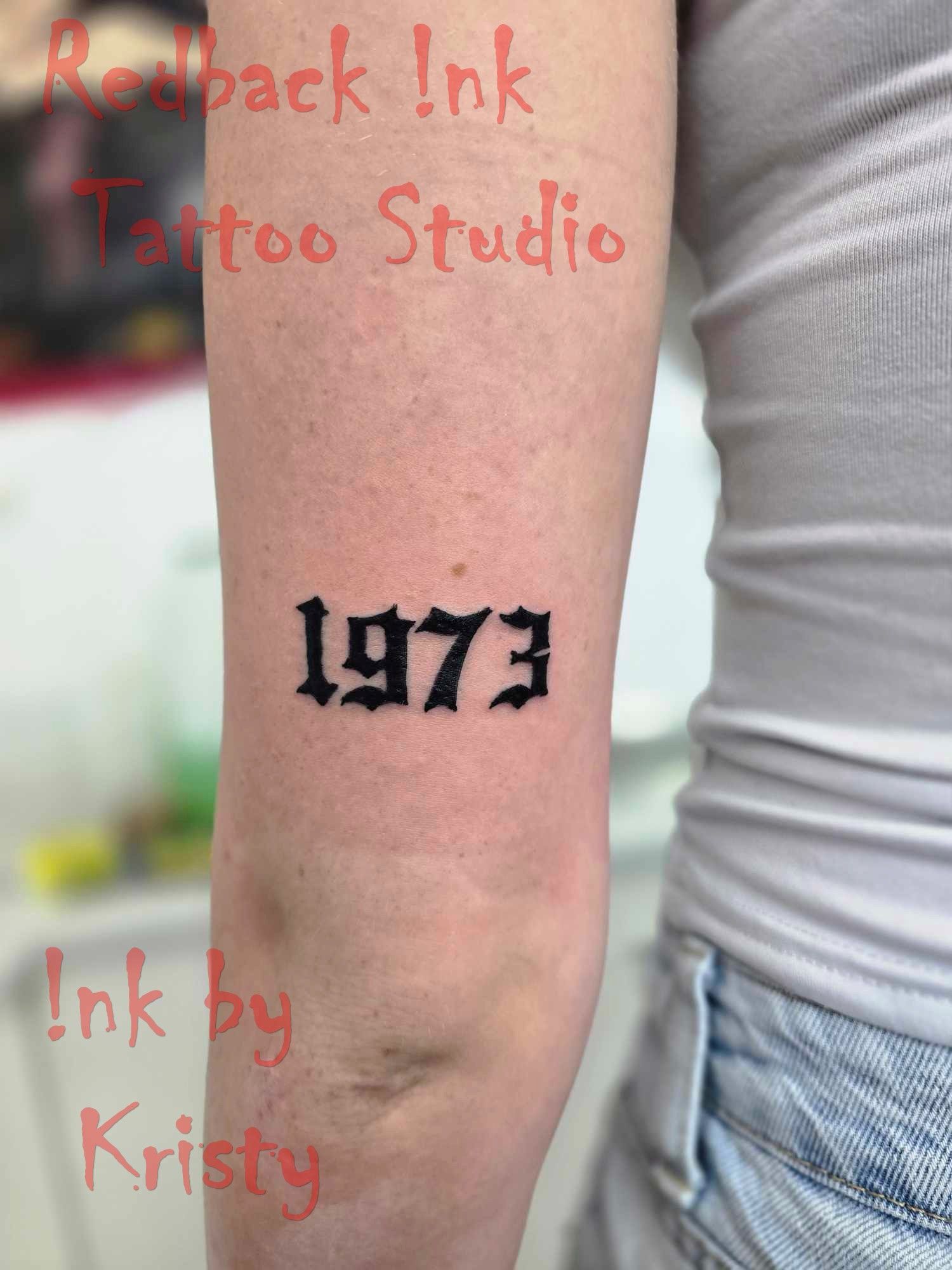 Black Tattoo of the Year 1973 on a Person's Arm — Redback !nk Tattoo Studio in Kawana, QLD
