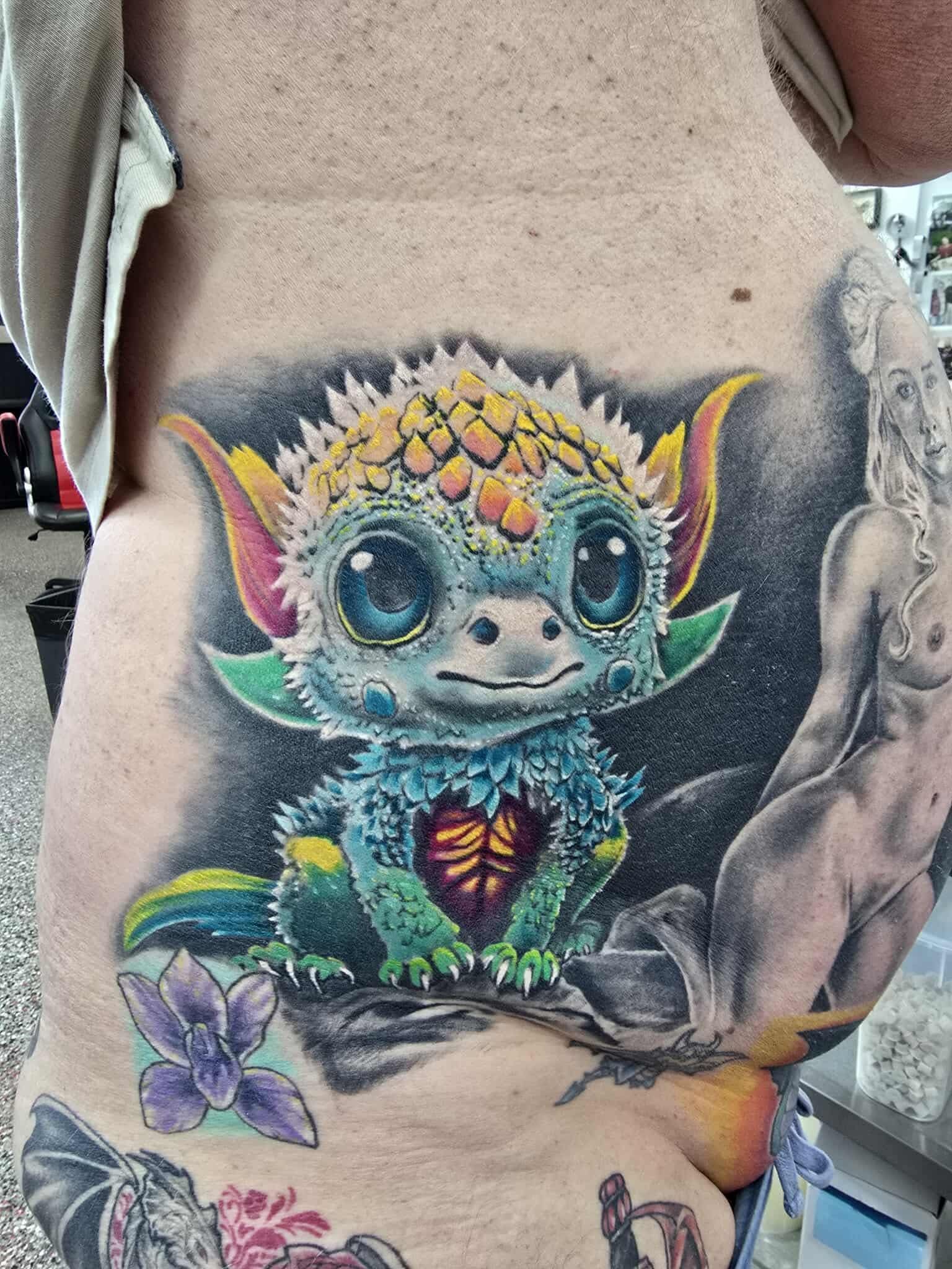 Tattoo of a cute, colorful dragon-like creature with large eyes, on a person's side with other tattoos.