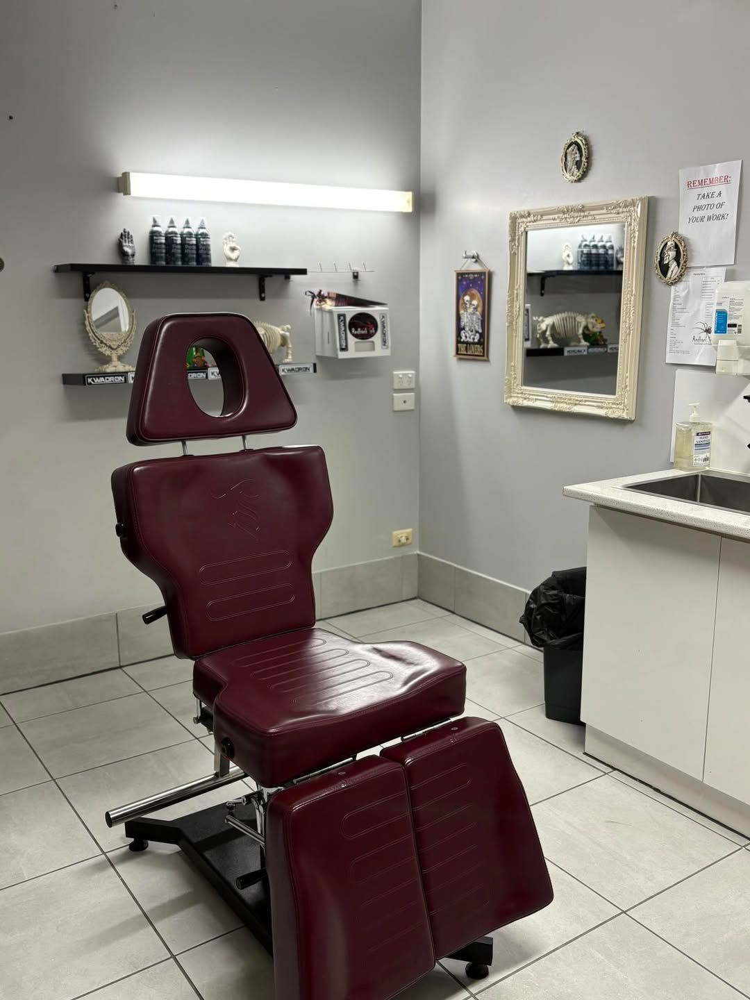 Tattoo artist's studio with a maroon chair, supplies, and mirror. White walls and tiled floor.