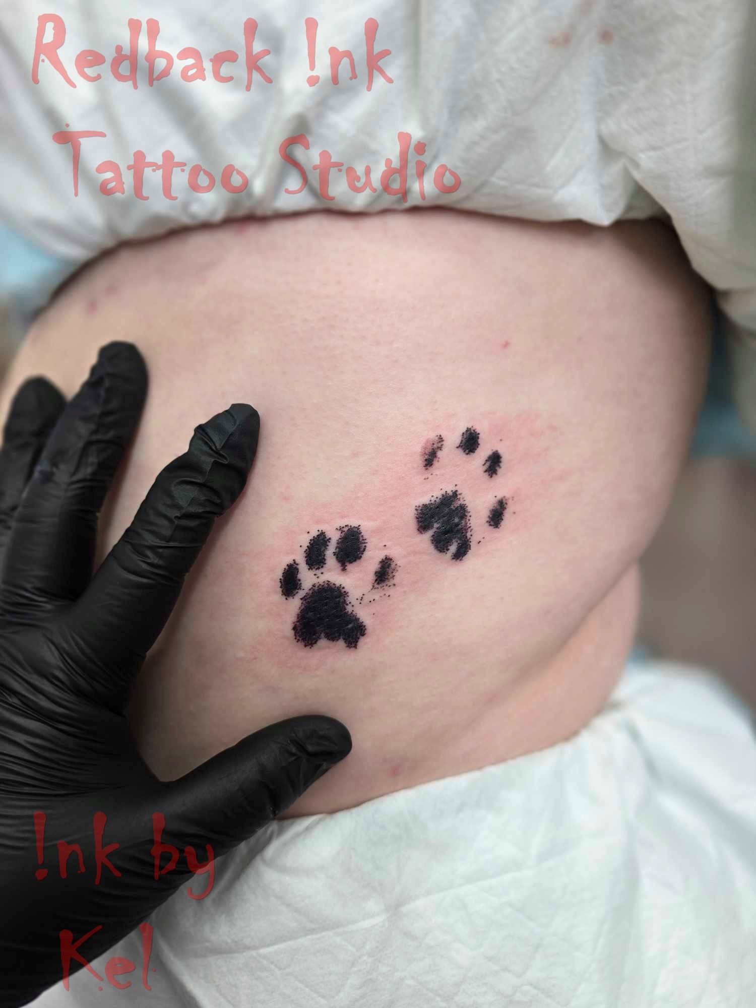 Two black paw print tattoos on light skin, held by a gloved hand. — Redback !nk Tattoo Studio in Kawana, QLD