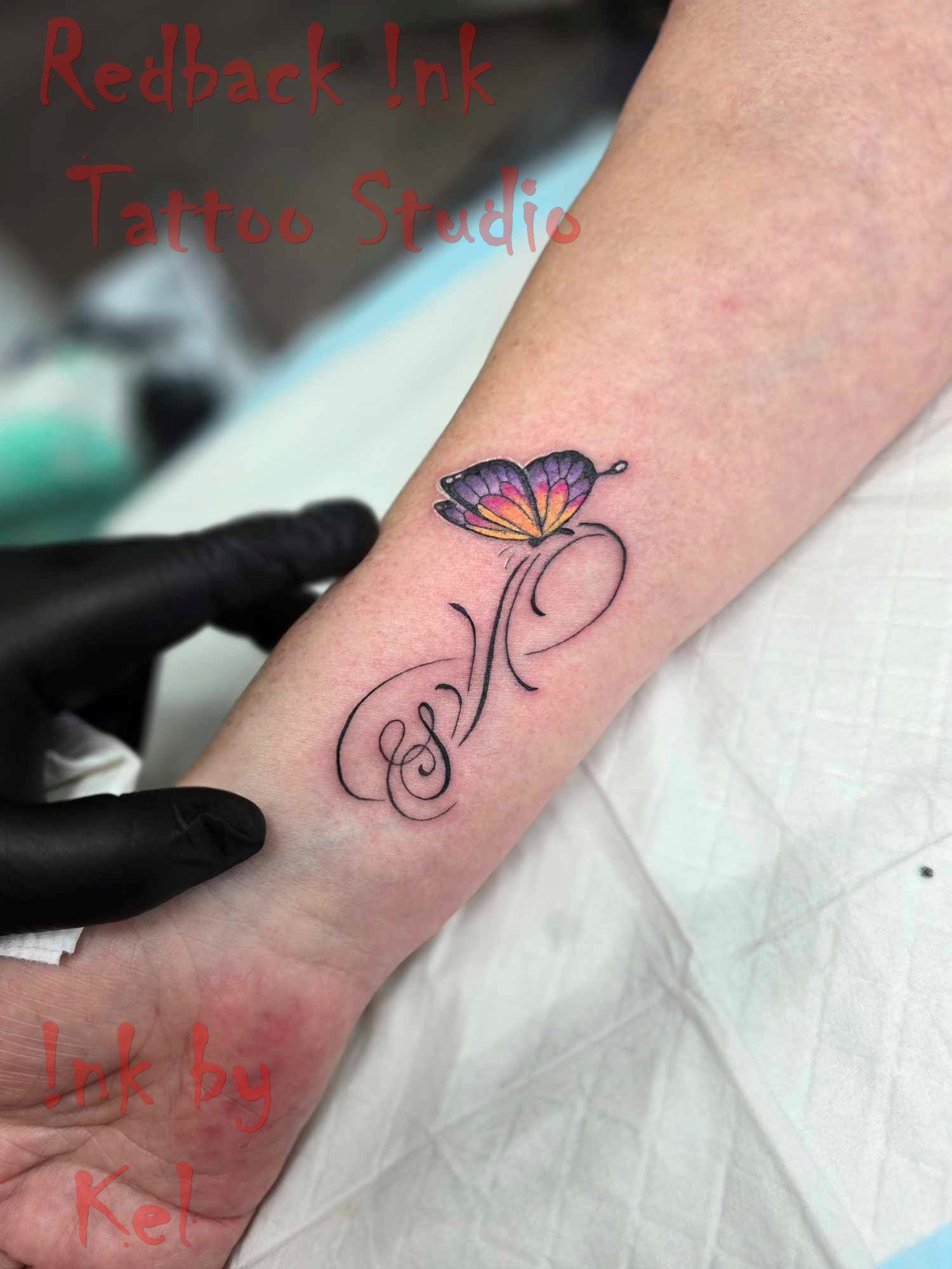 Tattoo of a colorful butterfly with initials on a wrist. Tattoo artist wearing gloves. — Redback !nk Tattoo Studio in Kawana, QLD