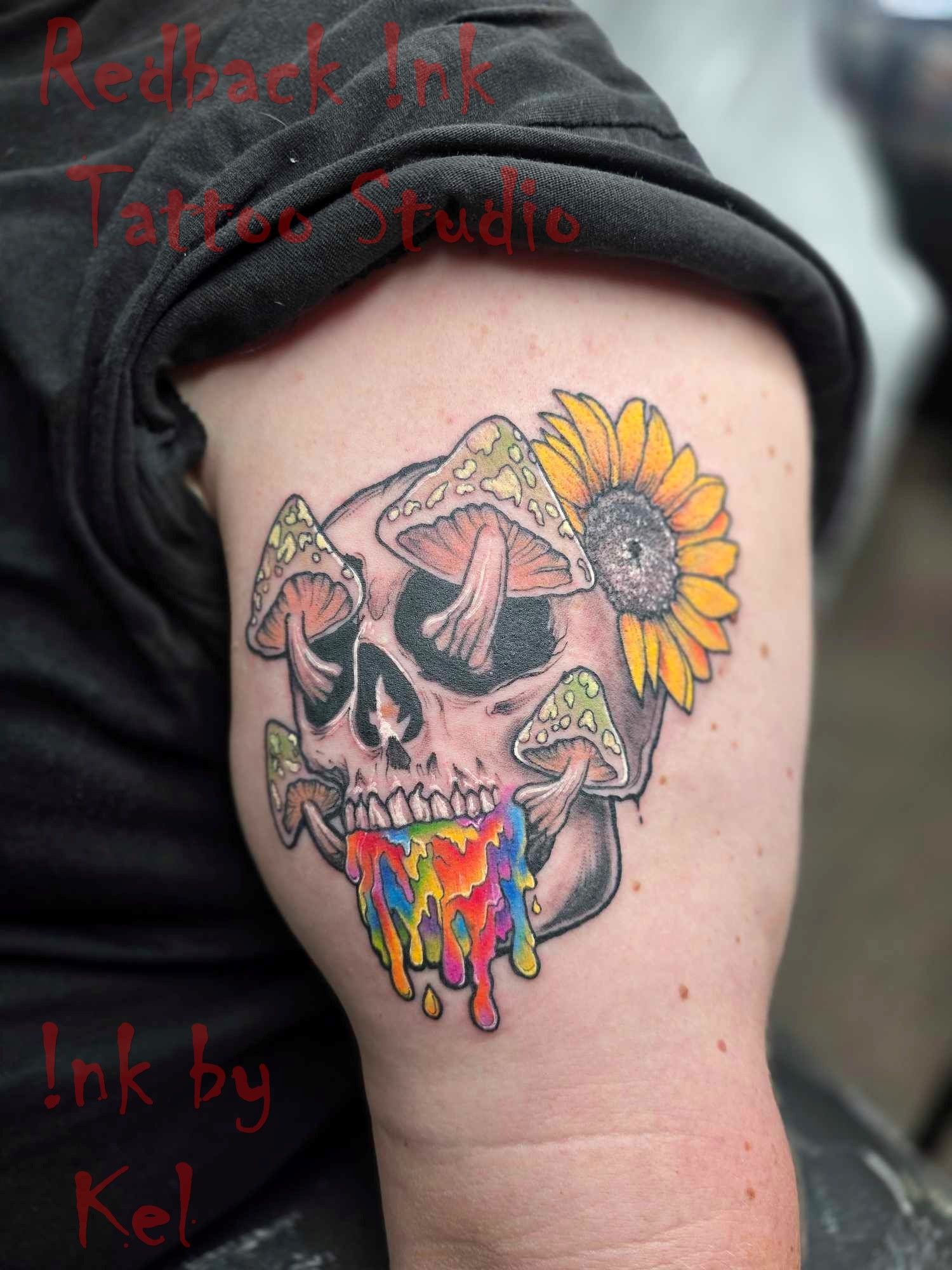 Skull tattoo with rainbow dripping from mouth, mushrooms, and sunflower on upper arm.