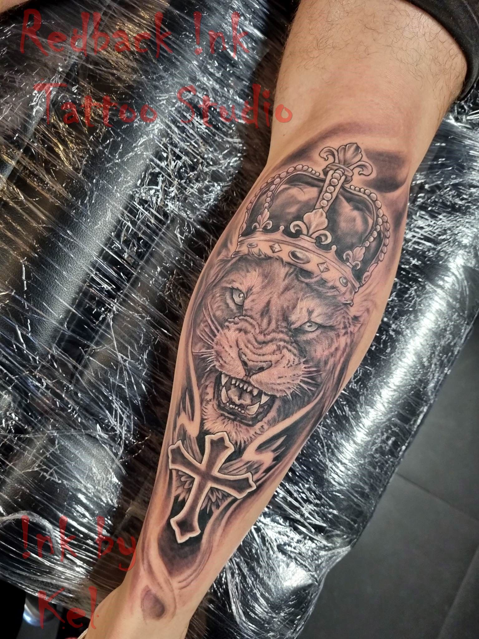 Lion's head with crown tattoo on arm; grayscale. A cross design is at the bottom.