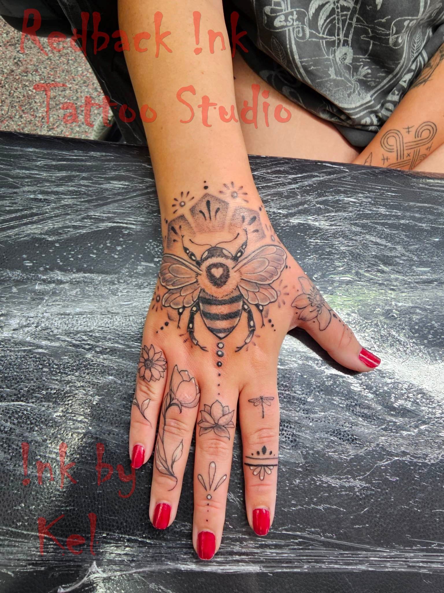 Hand tattoo of a bee with floral accents, red nails, on a person's hand, studio setting.
