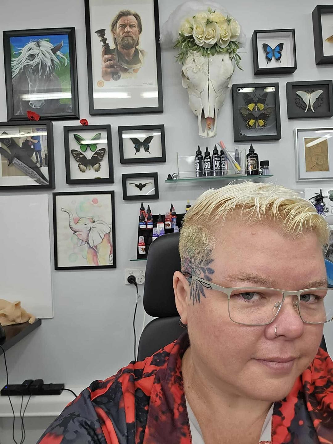 Blonde person with glasses and a flower tattoo, in front of a wall of art and tattoos.