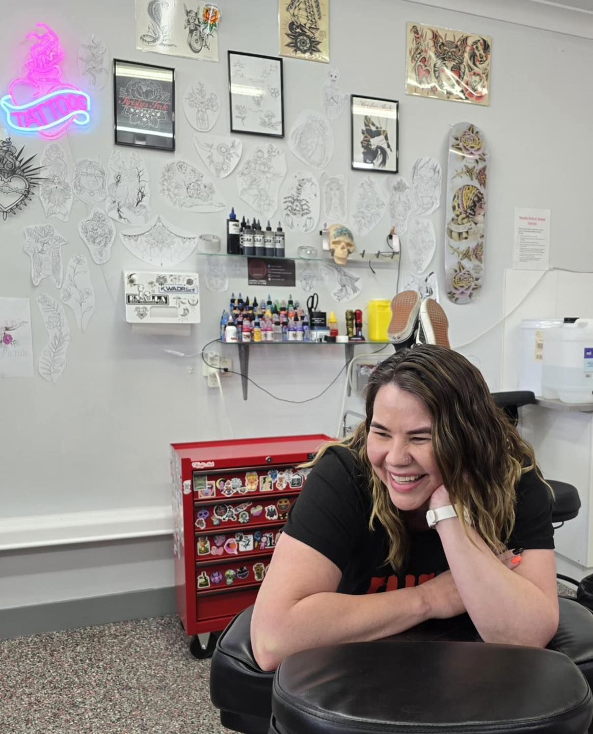 Woman laughing in a tattoo shop, leaning on chair. Grey walls with tattoo designs.