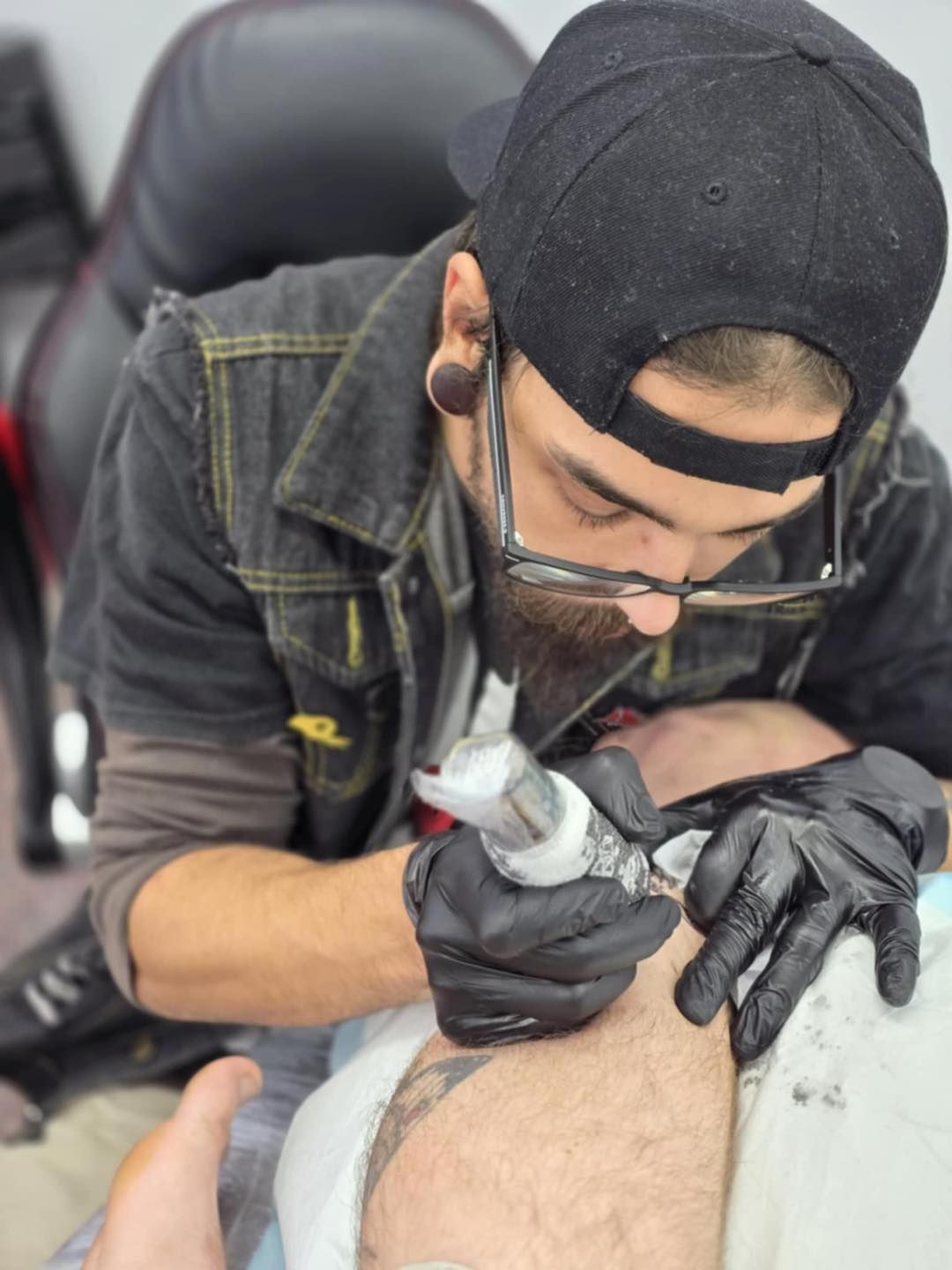Tattoo artist with black gloves and cap, working on a client's leg in a studio setting — Redback !nk Tattoo Studio in Kawana, QLD