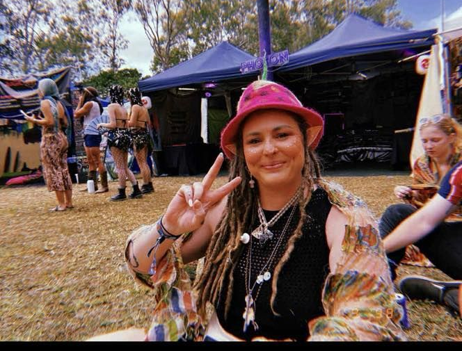 Person wearing a pink hat gives a peace sign, set outdoors with tents and trees — Redback !nk Tattoo Studio in Kawana, QLD