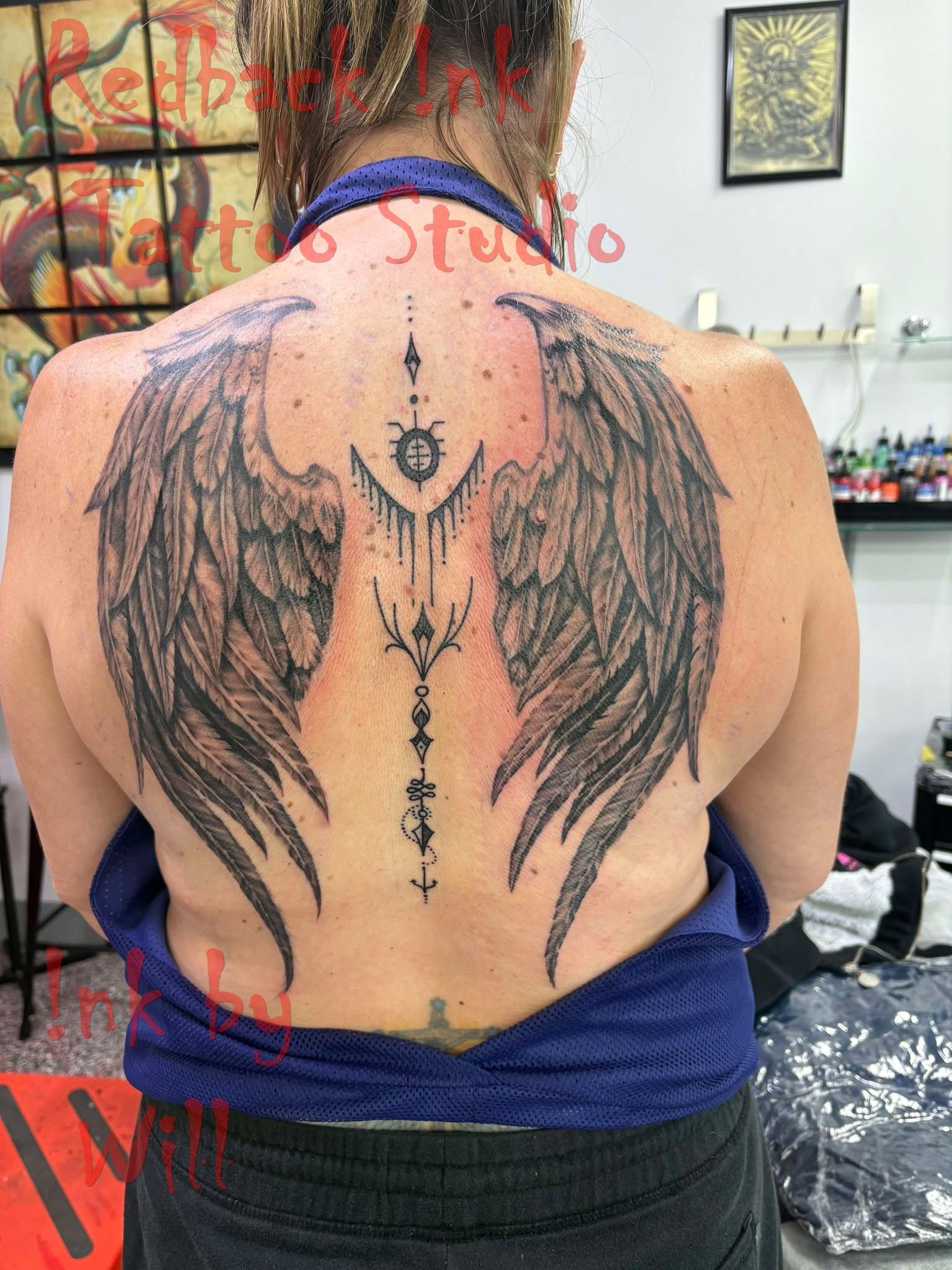 Woman's back with large black angel wings tattoo. Central design, dark ink, indoors.