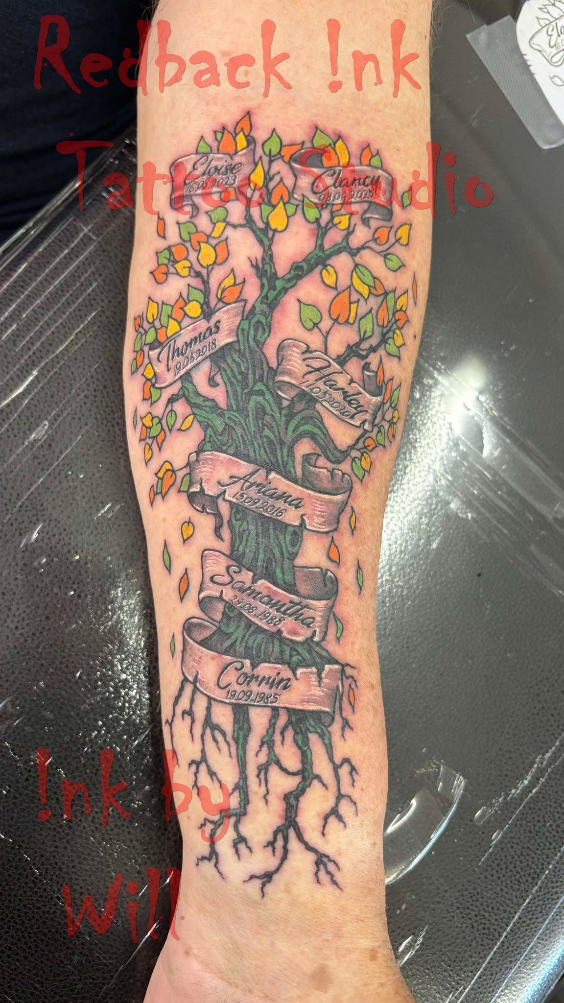Tattoo of a tree with colorful leaves, a banner, and roots on an arm.