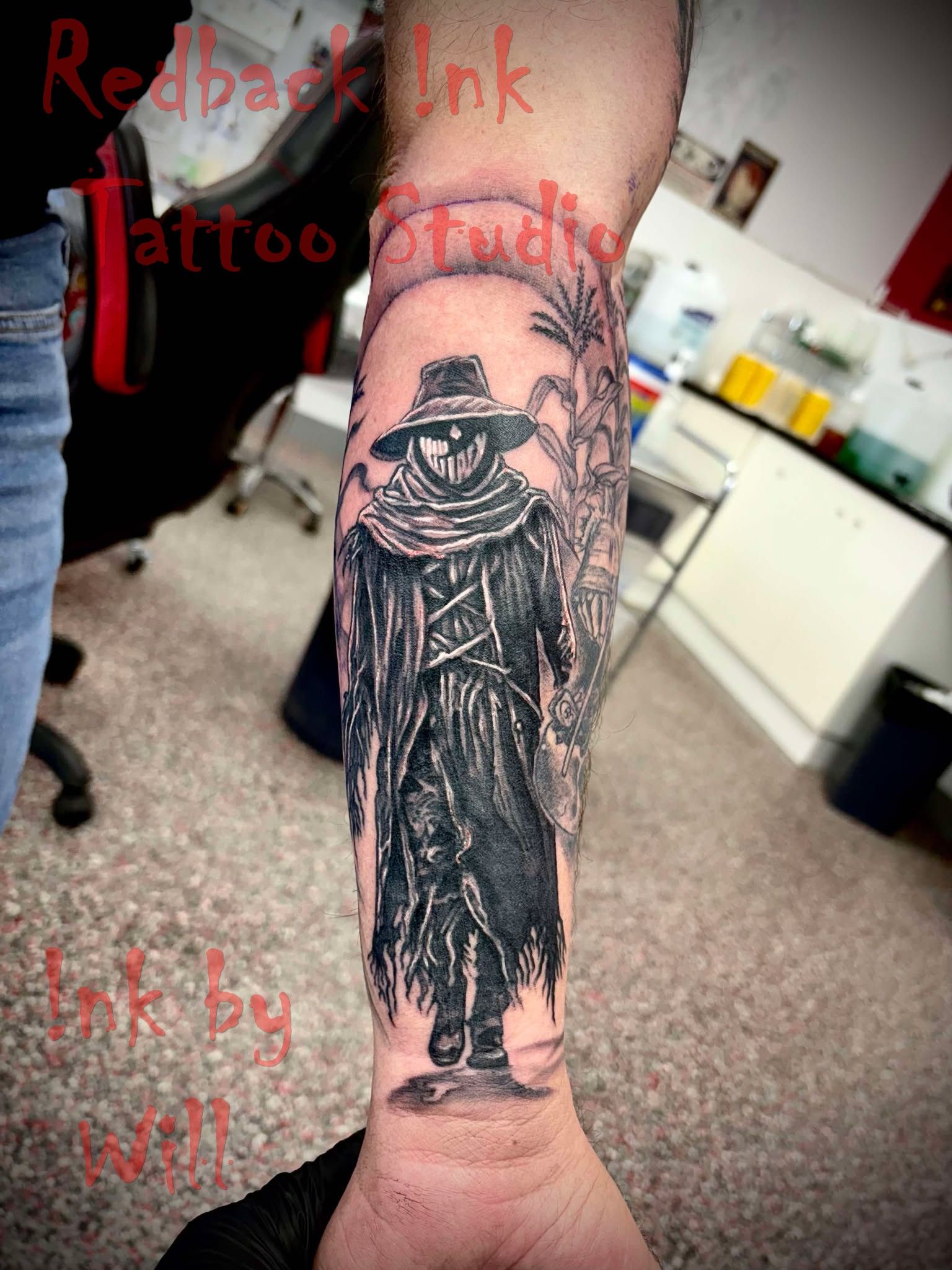 Black and gray forearm tattoo of a scarecrow wearing a hat and coat. The background is a circle with a blurred design.