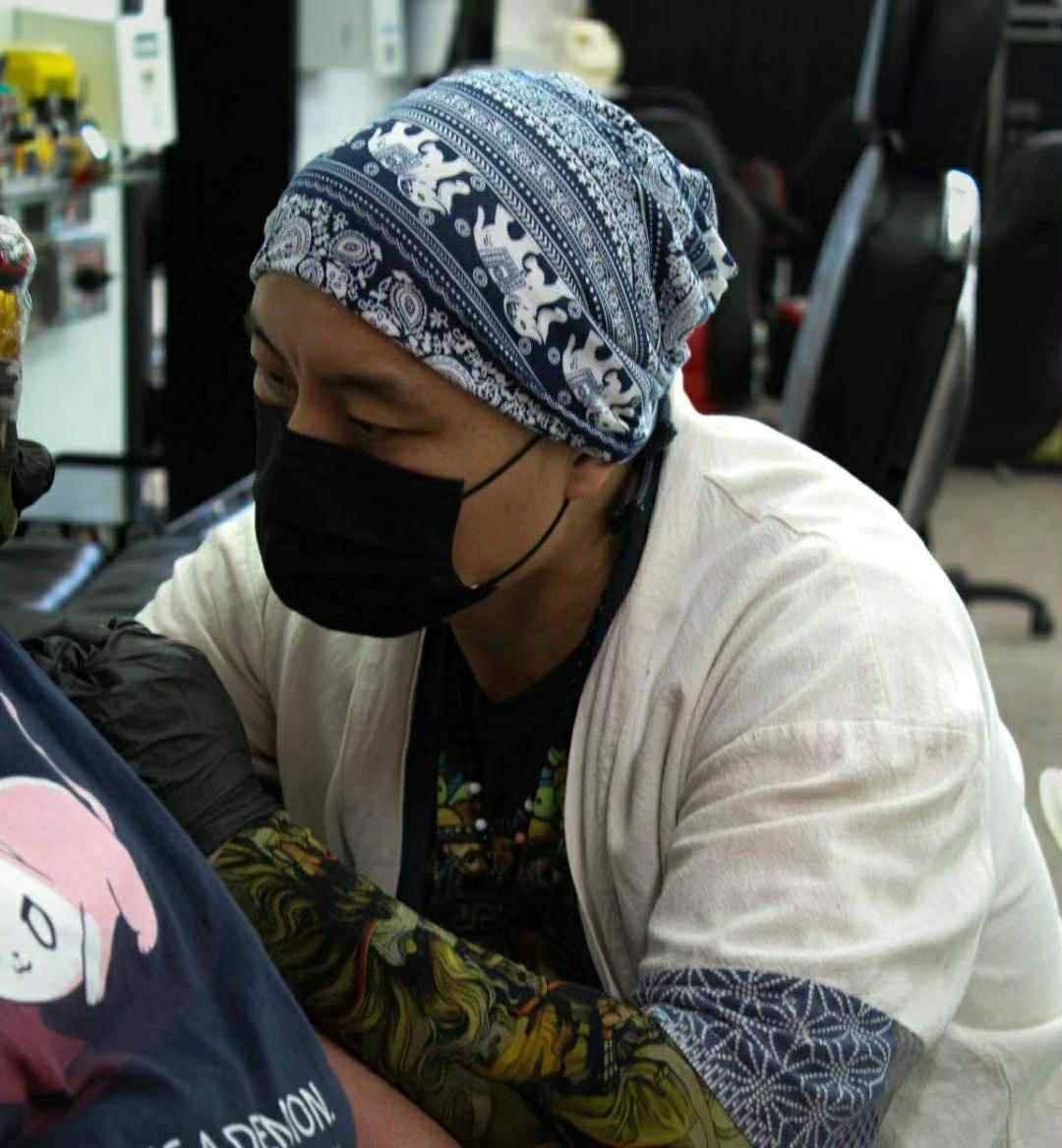 Tattoo artist wearing a mask and elephant-print headscarf, working on a client's arm.