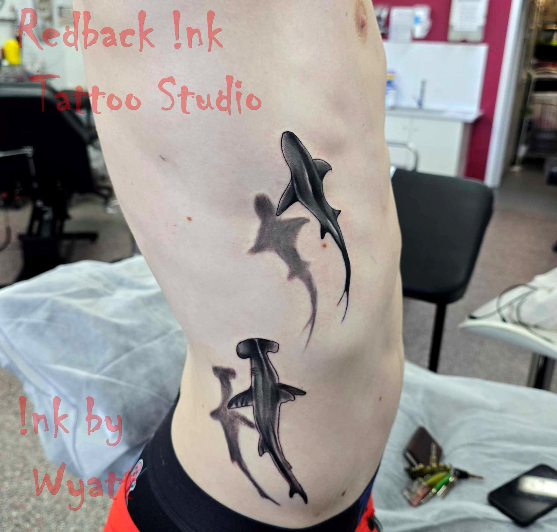 Tattoo of two sharks on a person's side.  One is a hammerhead, the other a regular shark.  Black ink. — Redback !nk Tattoo Studio in Kawana, QLD