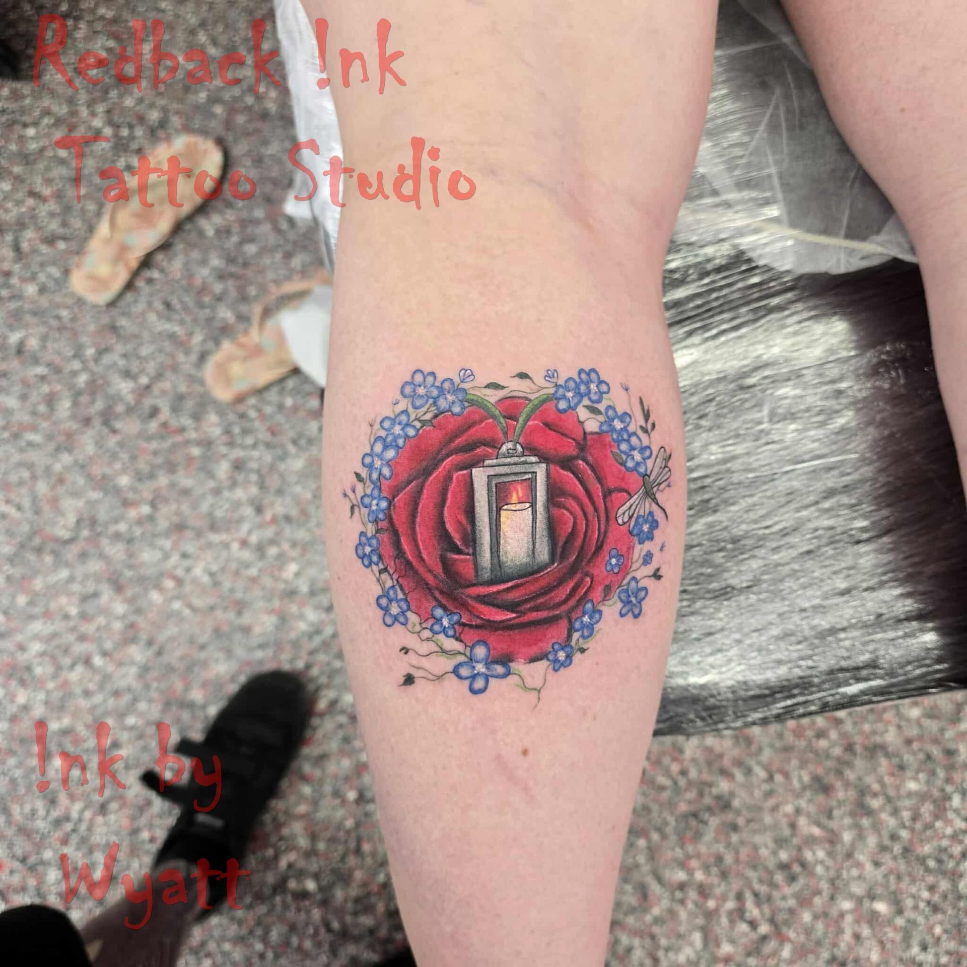 Tattoo of a red rose with a lantern holding a lit candle, surrounded by blue flowers, on a person's leg — Redback !nk Tattoo Studio in Kawana, QLD