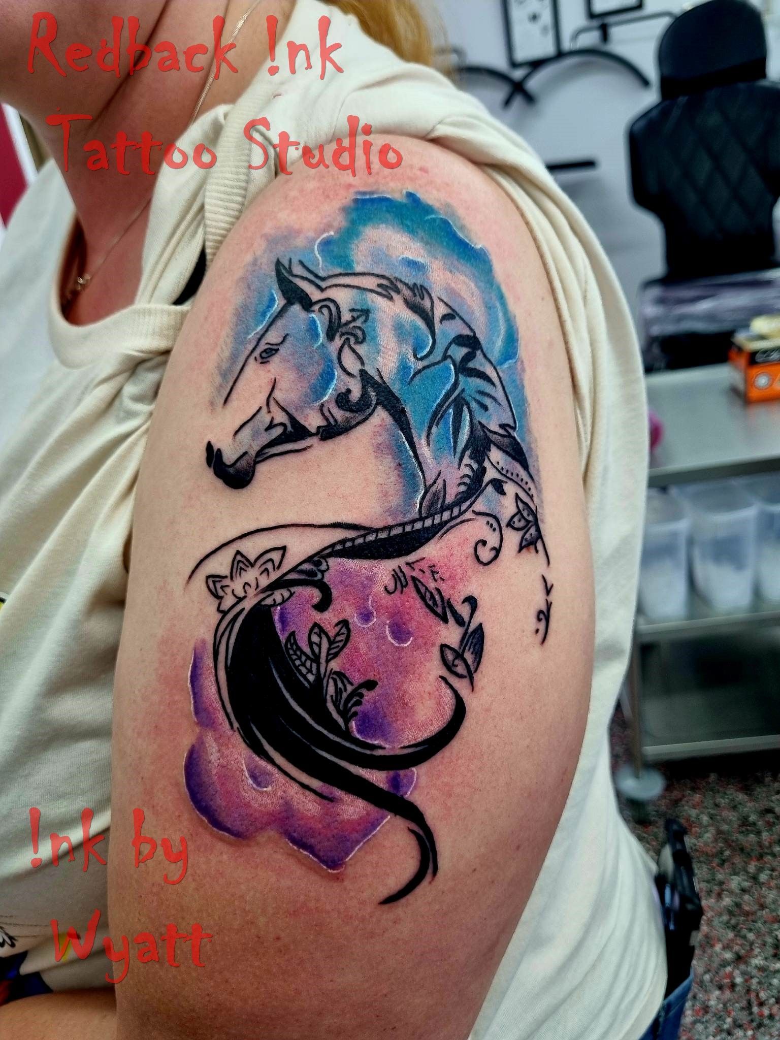 Tattoo of a horse head and body with purple and blue watercolor background on a person's upper arm. — Redback !nk Tattoo Studio in Kawana, QLD