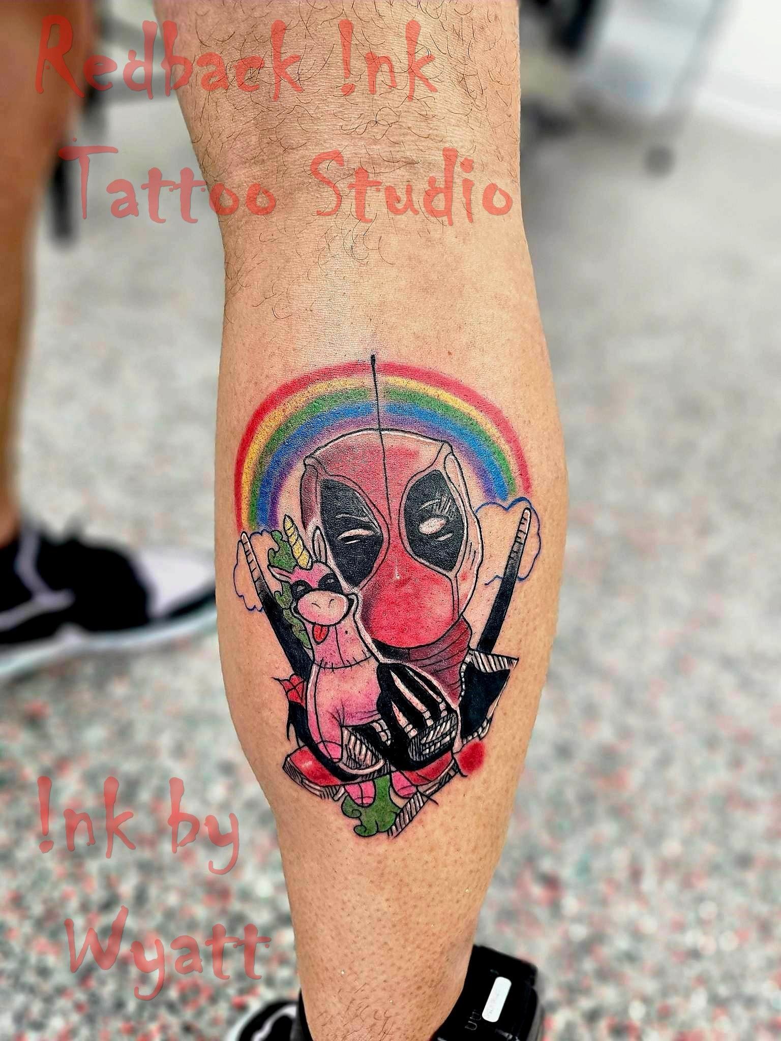 Deadpool tattoo with rainbow, unicorn, and swords on a leg. Colorful design with red, pink, and blue hues. — Redback !nk Tattoo Studio in Kawana, QLD