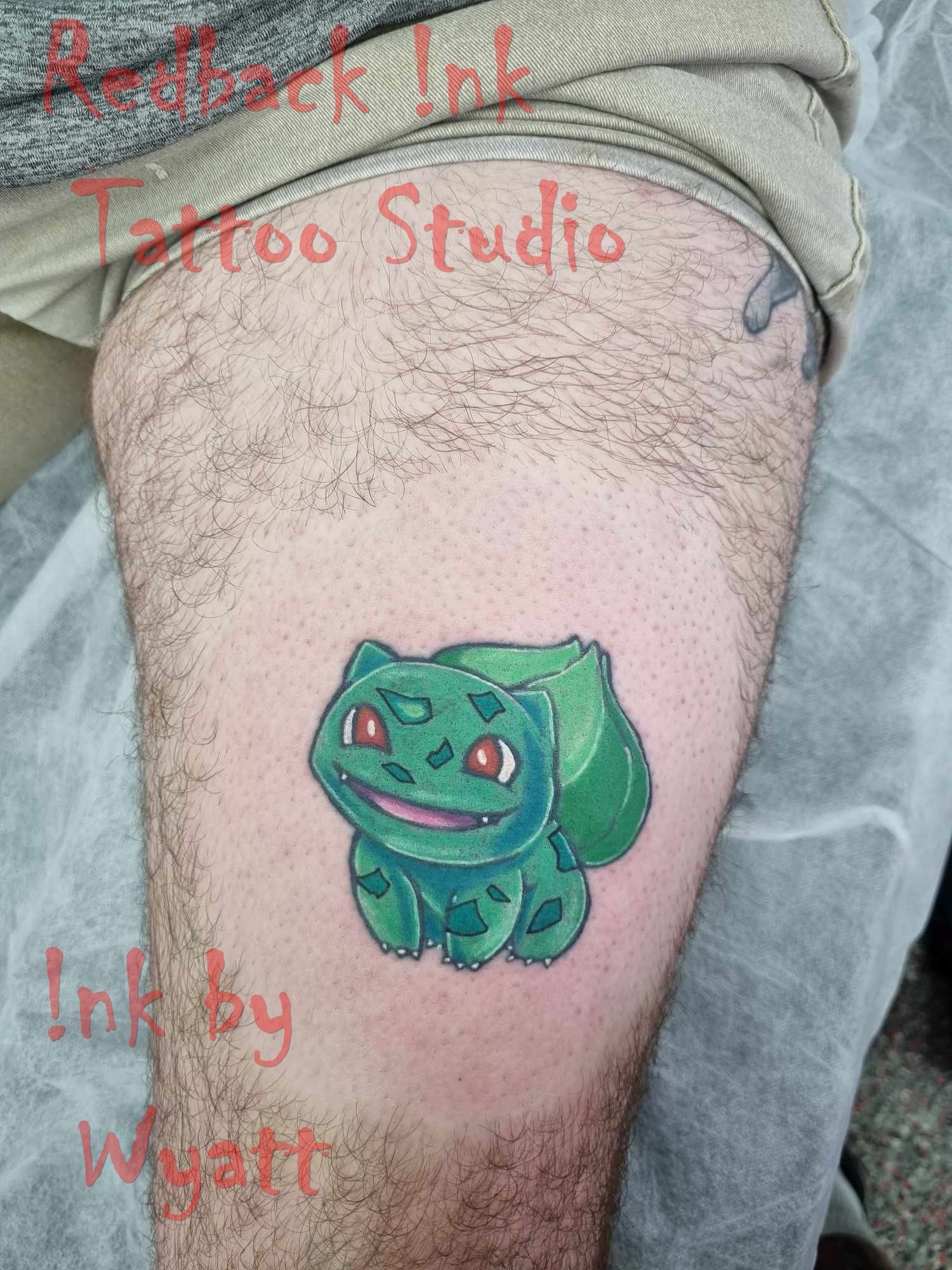 Bulbasaur tattoo on a leg. Green and blue Pokémon with red eyes, on a hairy thigh. Tattoo studio background. — Redback !nk Tattoo Studio in Kawana, QLD