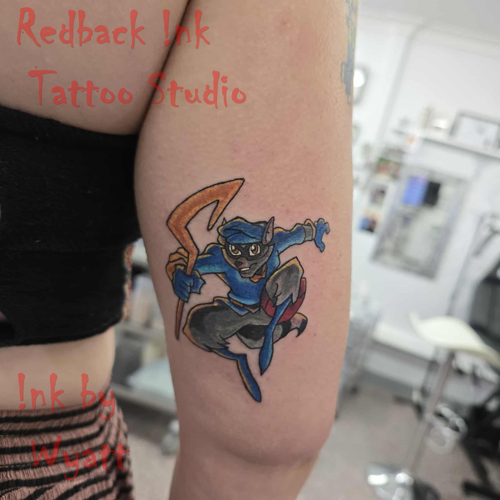 Tattoo of Sly Cooper in a dynamic pose, holding his cane. Blue, gray, and orange on an arm in a tattoo parlor — Redback !nk Tattoo Studio in Kawana, QLD