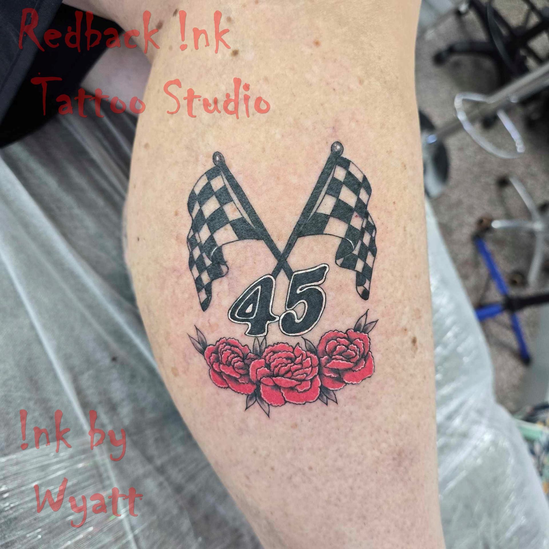 Tattoo of two crossed checkered flags, number 45, and red roses on a leg.