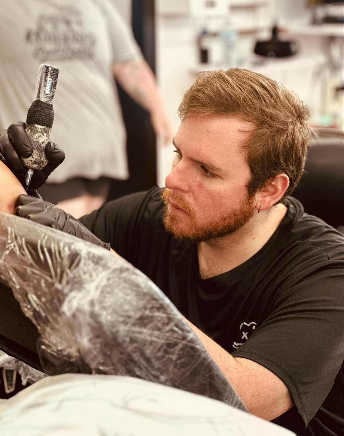 Tattoo artist focused, inking arm wrapped in plastic, wearing gloves. Shop setting.