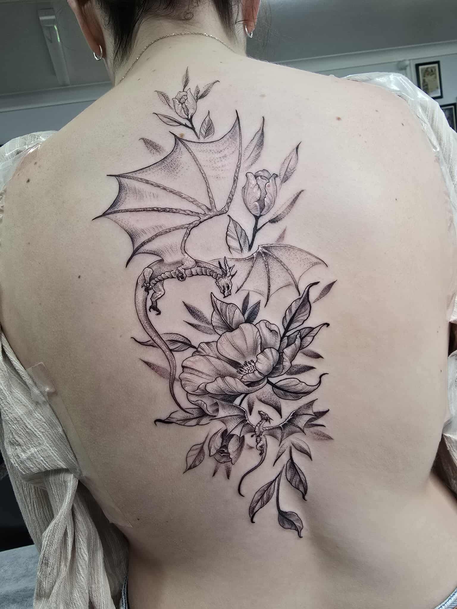 Tattoo on a back; dragon with wings entwined with flowers and leaves in black ink.