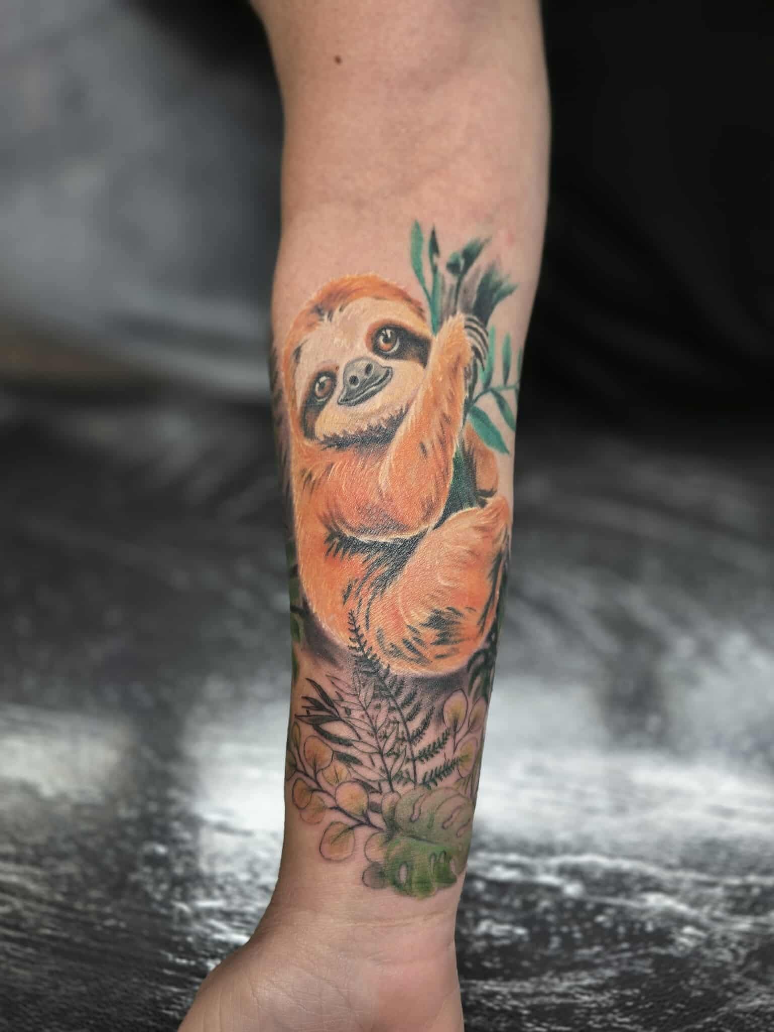 Sloth tattoo on arm. Orange-brown sloth hanging from leafy branch, surrounded by green and gray vegetation.