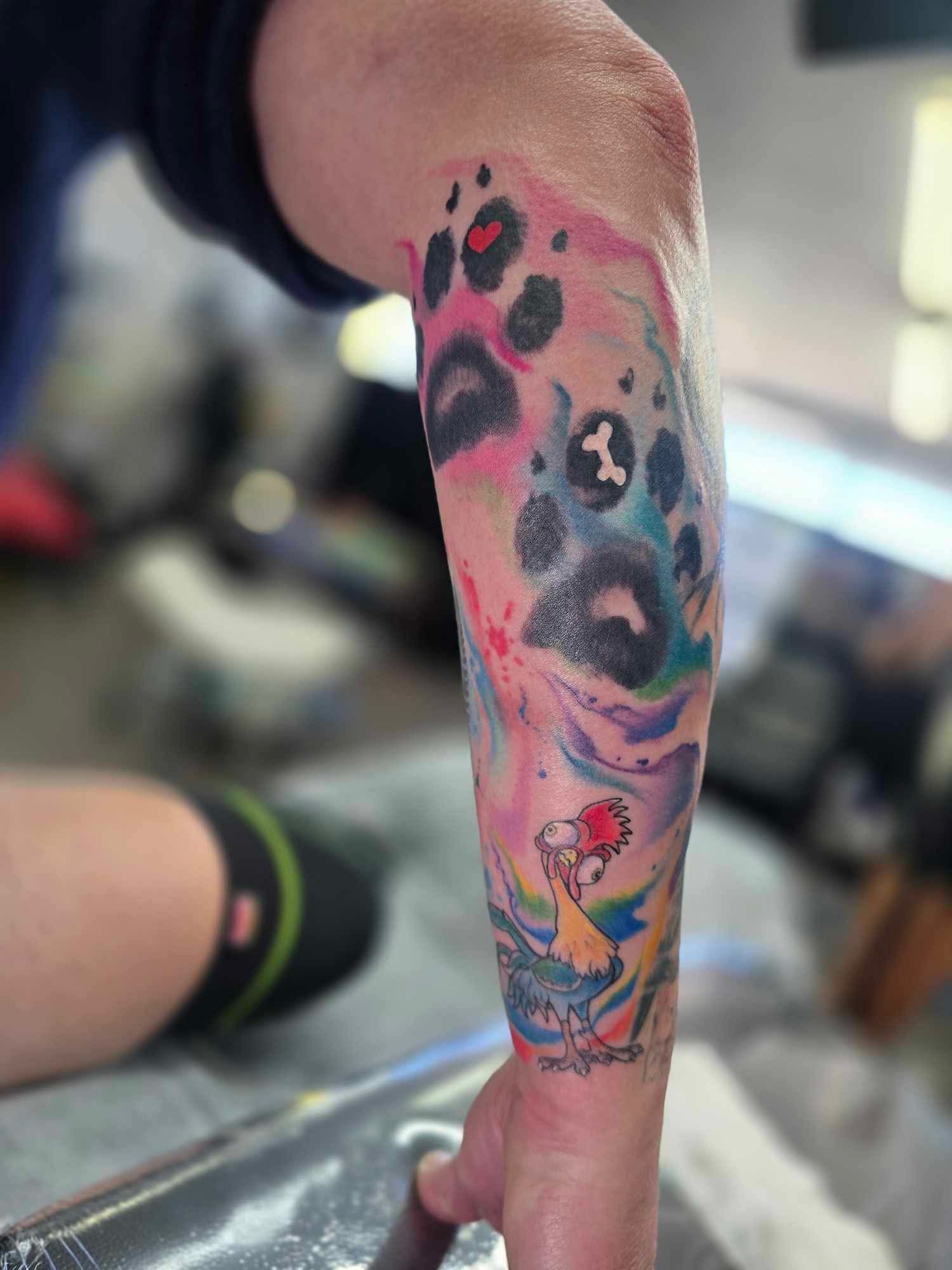 Colorful watercolor tattoo on forearm, features paw prints, abstract shapes, and bird.